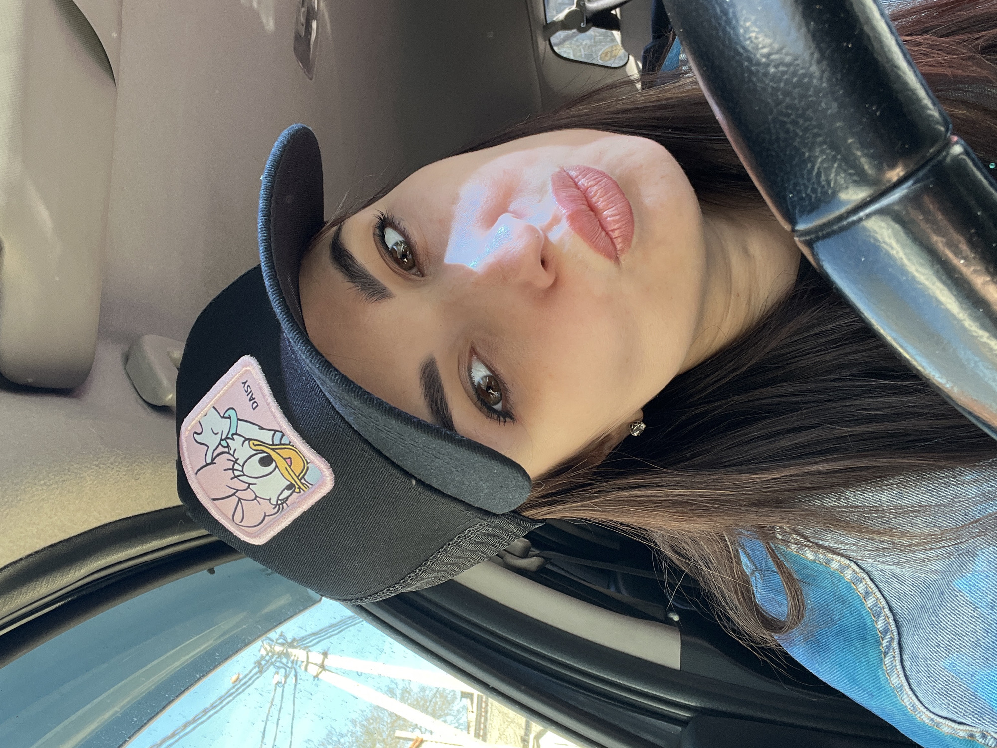 A person with brown hair and light skin is in a vehicle, wearing a black baseball cap adorned with a Daisy Duck patch. The person is positioned behind the steering wheel. The location is unknown.