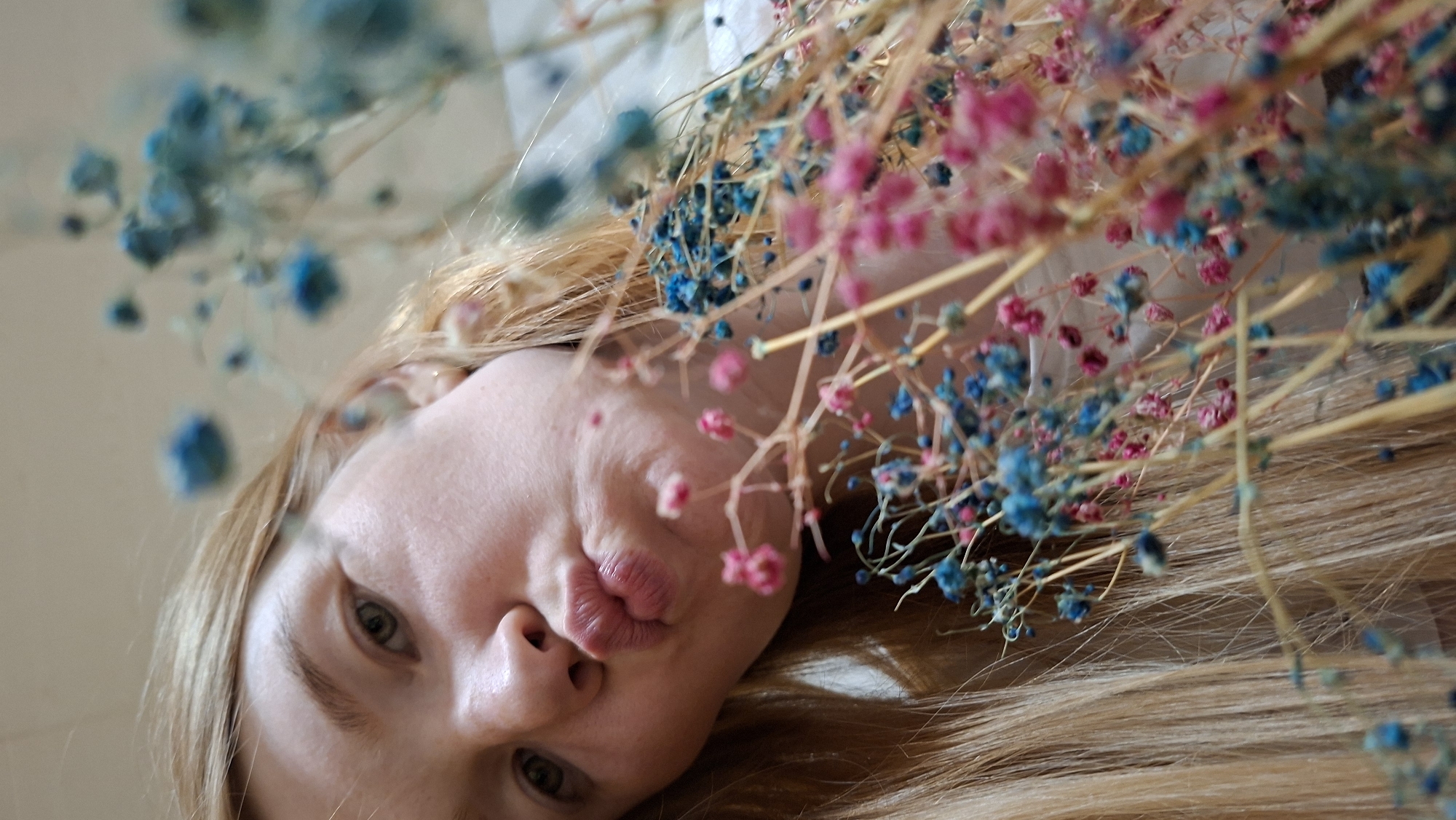 A close-up, low-angle shot captures a young woman with blonde hair puckering her lips as if blowing a kiss. She is positioned in the foreground, with a bouquet of dried flowers, featuring small pink and blue blossoms, obscuring part of her face and extending into the frame. The background is blurred and appears to be an indoor setting. The overall lighting is soft and natural. The location in the city of Большой Букор, Russia, is not discernible from the visual information present in the image.