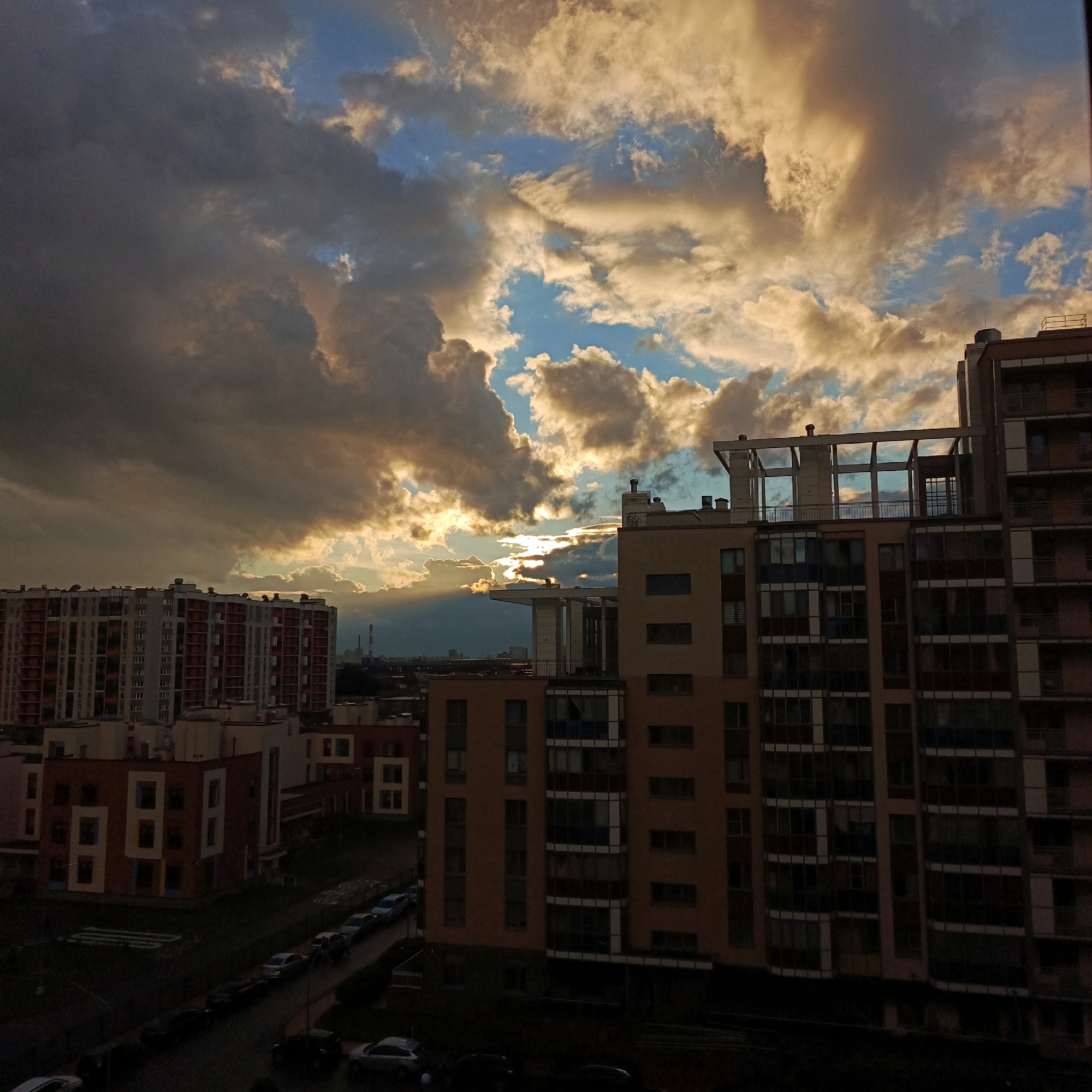 The image depicts a cityscape under a dramatic, cloudy sky, likely at dusk. In the foreground, several modern apartment buildings are visible, characterized by their varied architectural styles, modular designs, and numerous windows. The buildings are multi-story, with some featuring balconies and rooftop structures. Below the buildings, a street with parked cars and what appears to be a playground or recreational area can be observed.
The sky is a prominent element, filled with large, textured clouds that are illuminated by the setting sun, creating a contrast between dark, shadowed areas and brighter, golden patches. Rays of sunlight are piercing through gaps in the clouds, suggesting the sun is low on the horizon. In the distance, more buildings and possibly industrial structures, including a tall chimney, are faintly visible.
Based on the information that this image is from Bugry, Russia, the scene likely captures a residential development in this suburban area. The style of the buildings suggests a contemporary urban or semi-urban environment. The overall impression is one of an urban landscape experiencing the transition from day to night, with natural light playing a significant role in the atmosphere. There are no people clearly visible in the image.