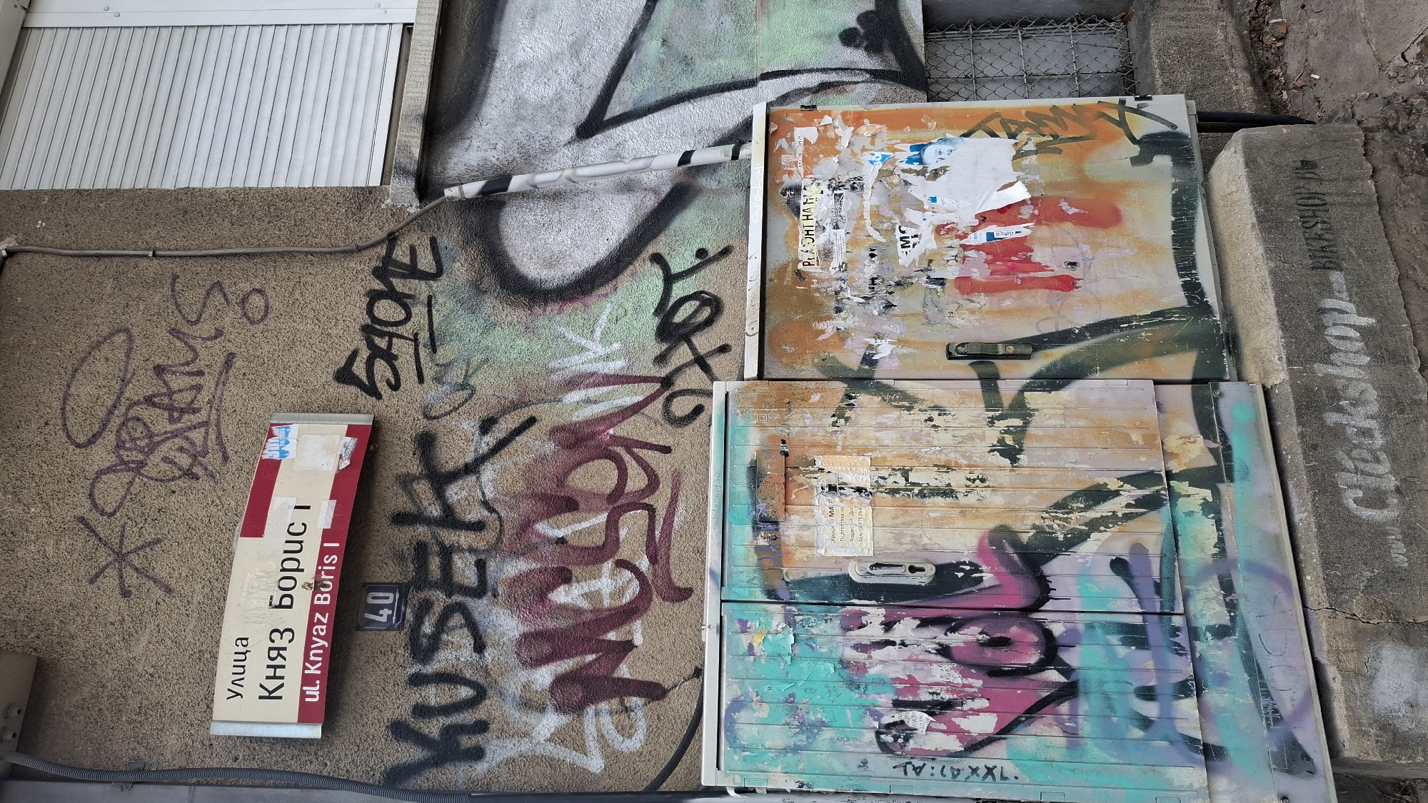 A street sign reads "Улица Княз Борис I" and "ul. Knyaz Boris I" in Sofia, Bulgaria. Below the street sign, the number 40 is displayed on a black plaque. The wall behind the sign is covered in graffiti. In front of the wall are two utility boxes, also covered in graffiti, with a concrete base in front of them. The concrete base has "www.Clockshop - BIKESHOP.BG" spray-painted on it.
