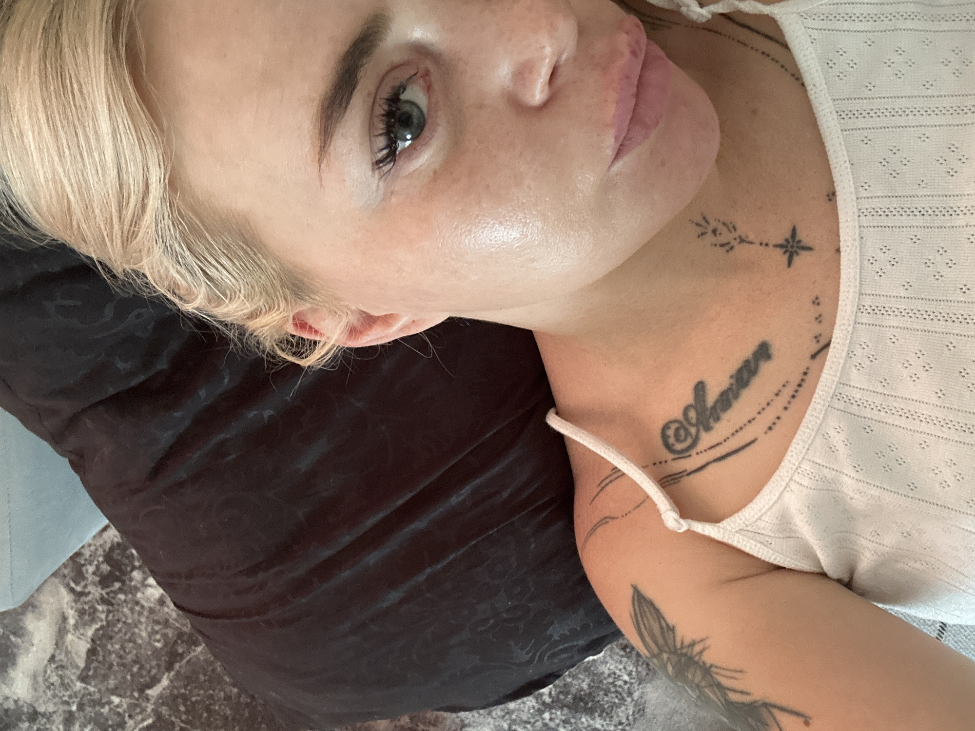 An extreme close-up, overhead shot focuses on the right side of a woman's face as she lies on a dark, crumpled fabric. Her blonde hair is pulled back, and her right eye, with dark lashes and a green iris, is partially visible. Her skin appears luminous and slightly oily, with some freckles visible on her cheek. A portion of her shoulder and chest are also in view, adorned with tattoos. She wears a white, ribbed tank top with thin straps. The background consists of dark fabric and a hint of a gray, marble-like surface. The image has a shallow depth of field, blurring the background and highlighting the woman's features.