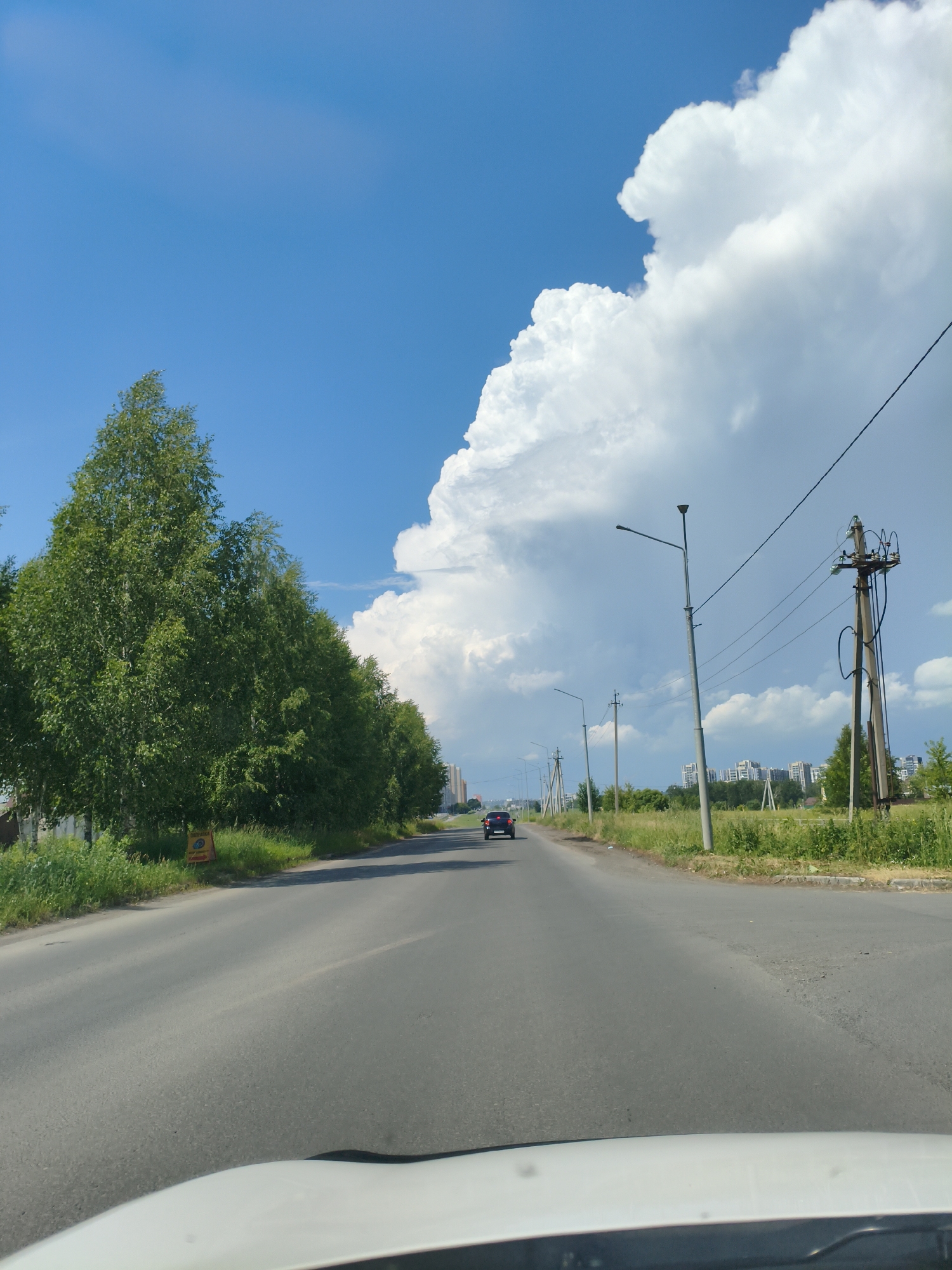 This is a photograph taken from a vehicle, looking forward along an asphalt road. On the left side of the road is a dense line of green trees, possibly birch trees, with a bright yellow sign visible amongst them. The right side of the road features a grassy verge with several utility poles and power lines. In the distance, several modern apartment buildings are visible, suggesting an urban or suburban setting. The sky is a bright blue, with a large, dramatic white cumulus cloud dominating the upper right portion of the frame. A single dark-colored car is driving away from the camera in the middle of the road. The overall impression is a sunny day with a sense of forward motion. This scene is consistent with locations in or around Металлплощадка, Russia, given the style of architecture and the presence of power infrastructure.