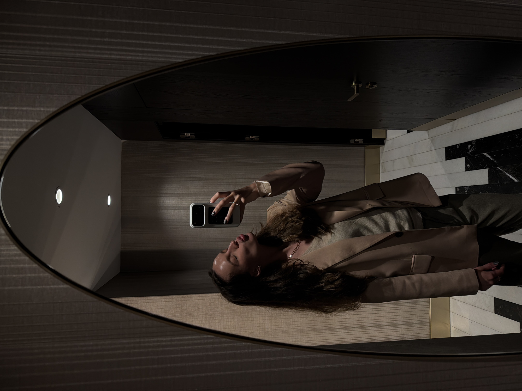 The image shows the reflection in an oval mirror of a woman taking a selfie. She is wearing a beige blazer and holding a smartphone. The background includes a wall with two recessed lights, a wood-paneled surface, and a marble-tiled wall. The location may be a bathroom or dressing room. Based on the décor style of the room, it can be inferred that the location may be in Budva, Montenegro.