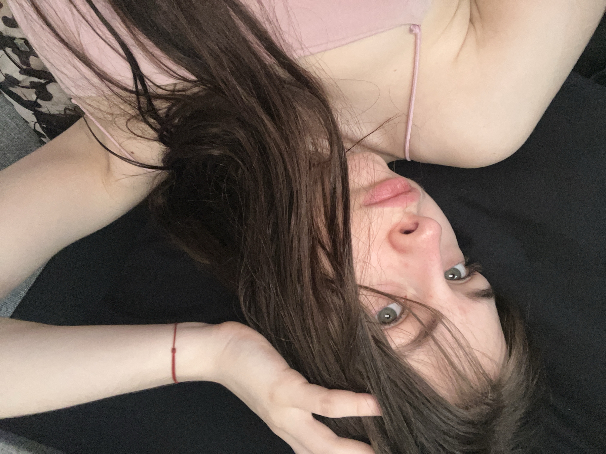 A woman with long, dark hair is lying on a dark surface, possibly a bed or couch. She is wearing a light pink tank top. Her face is in the foreground, with her eyes looking directly at the camera. She has fair skin and her hair is partially covering her face. A red string bracelet is visible on her wrist. The background is blurred and consists of a dark, indistinct surface and a patterned section to the left. The image lacks information to connect it to Saint Petersburg, Russia.