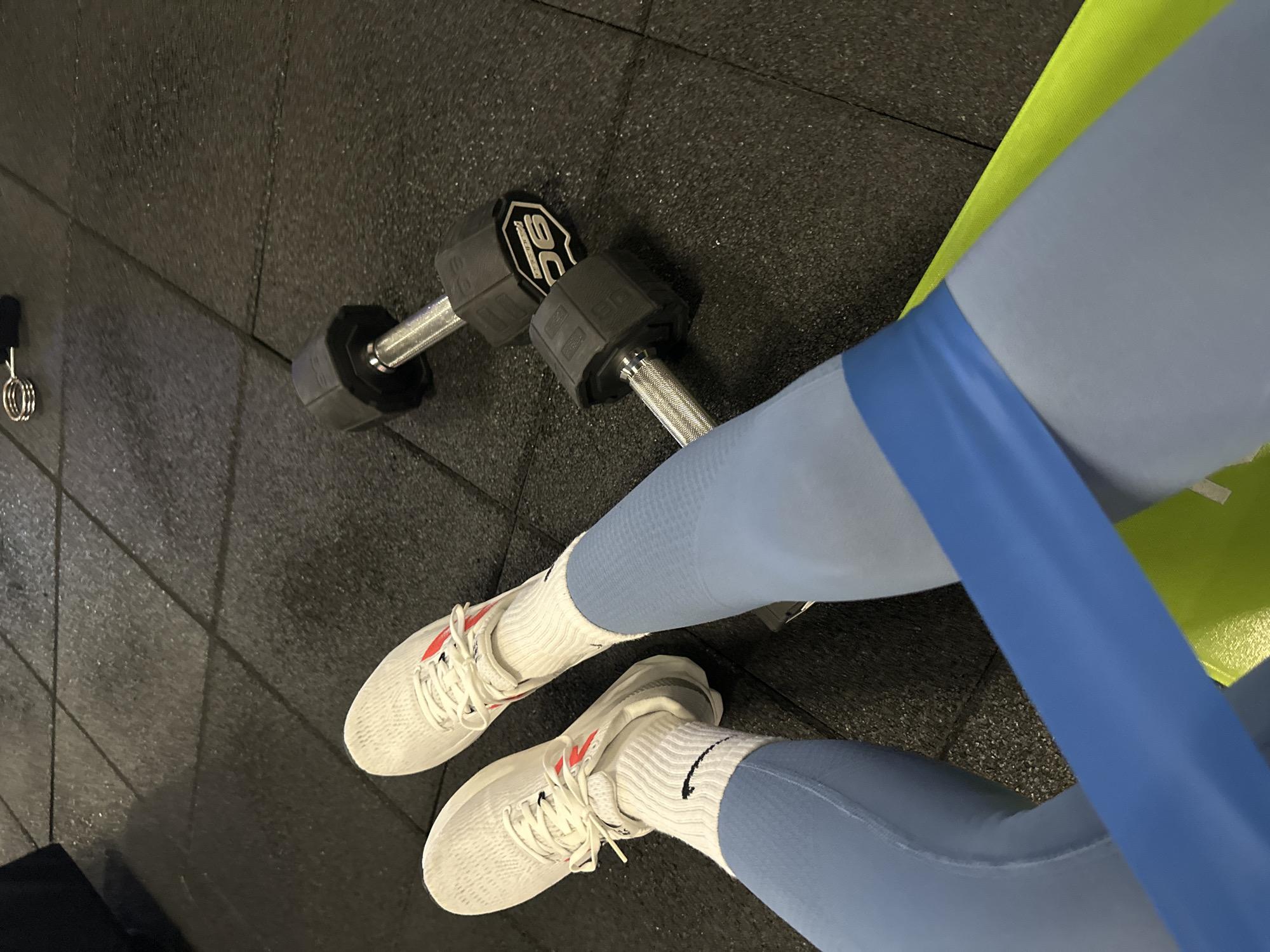 A top-down, medium shot depicts a pair of white tennis shoes with white socks, light blue workout pants with a blue band, a pair of black dumbbells, and a green yoga mat, all lying on a dark grey rubberized floor. The shoes are positioned at the bottom of the frame, angled slightly to the right. The dumbbell is located at the top left, while the workout pants are positioned in the center and angled to the upper right. The yoga mat is peeking from the pants. The location is Unknown, Unknown.