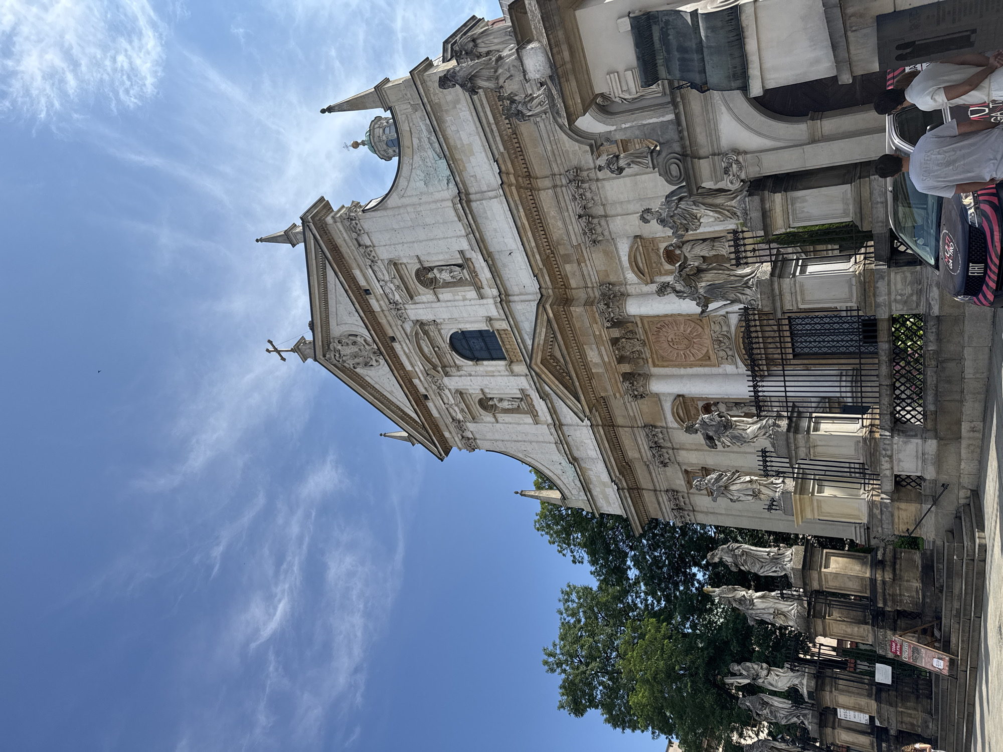 The image shows the Church of Saints Peter and Paul in Krakow, Poland, a Baroque Roman Catholic church. The facade of the church is white stone, adorned with numerous statues of saints and religious figures. The sky is blue with wispy white clouds. In front of the church, a line of statues on pedestals stands behind a black iron fence. To the left of the church, a large green tree is visible. In the foreground, a dark colored Fiat car with pink stripes and a logo that reads "Multi Gastro" is parked. Two people, a man and a woman, are standing near the car, facing away from the camera. Another person, a man, is also standing near the car, facing away from the camera. In front of the statues on the left, a wooden A-frame sign with text is visible. The overall scene depicts a sunny day outside a prominent religious landmark.
