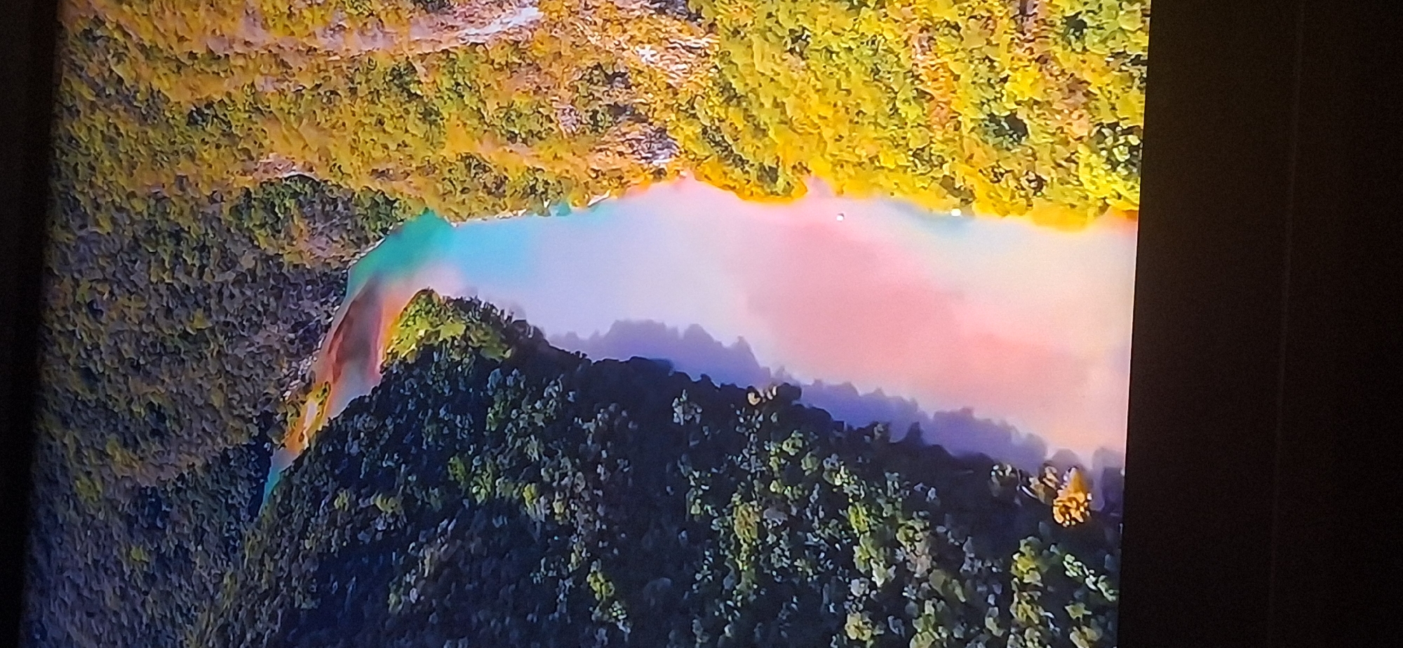 The image is an aerial view of a river flowing through a canyon. The river displays a gradient of colors, from turquoise at the top of the image, transitioning through pale pink and white, and then to a more muted blue further down. The canyon walls are covered in dense green and yellow foliage. The scene is illuminated by natural light, casting shadows that highlight the contours of the landscape. There are no visible people or man-made structures in the immediate vicinity. The image does not provide any information about the city of Bila Tserkva, Ukraine, or any connection to it.