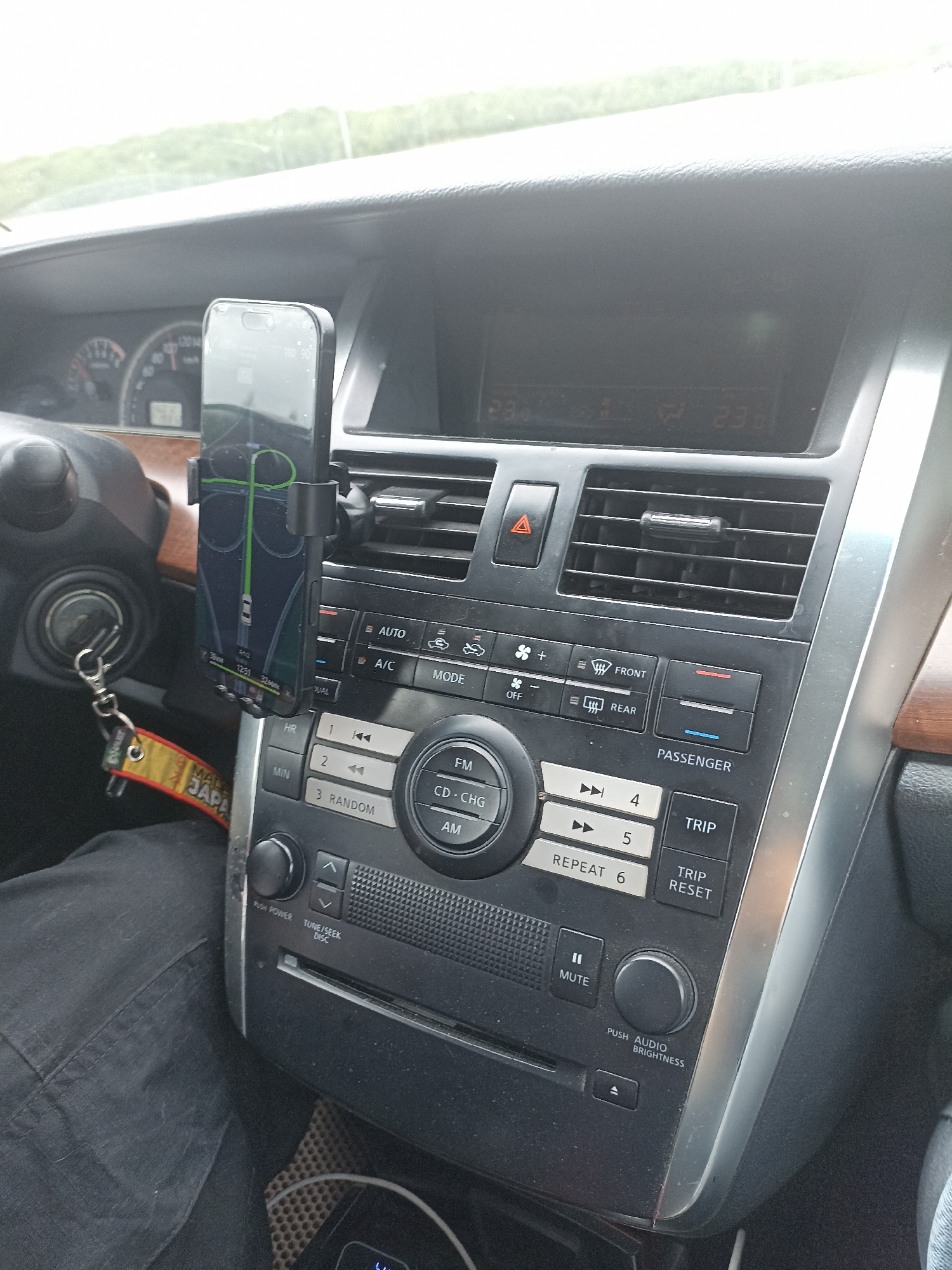 This media file is a close-up photograph taken inside a car, focusing on the central dashboard area. The image captures a smartphone mounted on a car mount, displaying a navigation app with a route highlighted in green. The phone screen is partially reflective, showing some smudges.
To the left of the phone, a portion of the steering wheel and ignition are visible. A keychain with a yellow and red tag featuring Japanese text ("MALAYSIA JEPANG" appears to be visible from the crops) hangs from the ignition. The car's dashboard features various controls, including buttons for climate control (AUTO, A/C, MODE), radio functions (FM, AM, CD-CHG), and trip information (TRIP, TRIP RESET). There are also several vents and a display screen showing digital readouts, seemingly indicating temperatures of "23.0" and "23.6". The interior of the car is dark, suggesting it might be daytime with overcast skies, as indicated by the bright, diffused light coming from outside, and the lack of harsh shadows. The overall scene is a snapshot of a car's interior, likely during a journey. No people are clearly visible in the frame, though the presence of a driver's leg covered in black fabric suggests someone is operating the vehicle. The car appears to be parked or stopped, given the focus on the dashboard and navigation system.
