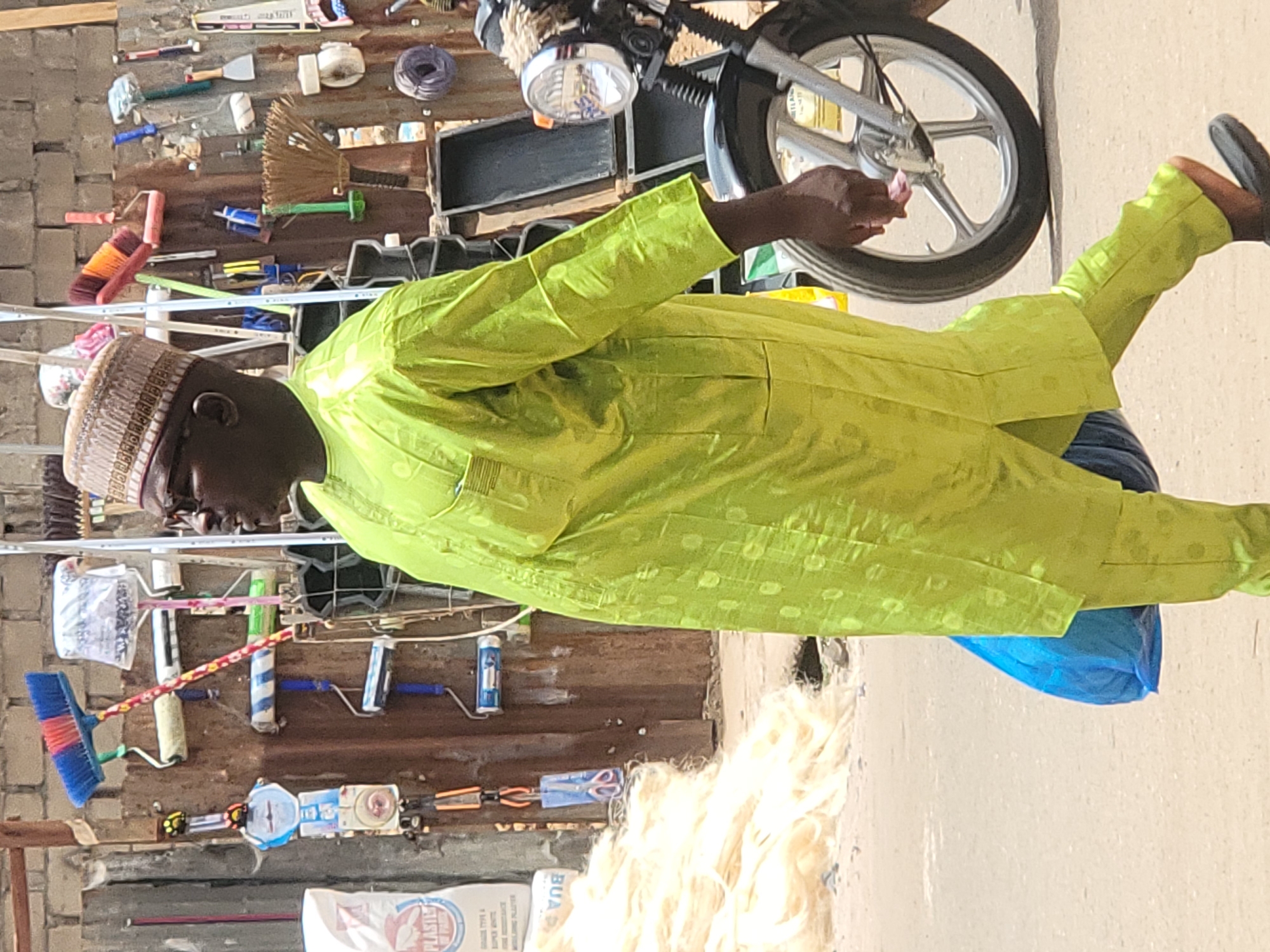 An outdoor, brightly lit image captures a man standing next to a street stall in what appears to be Maiburiji, Nigeria. The man, of medium complexion, wears a vibrant lime green traditional outfit (likely a kaftan or agbada) with subtle patterned texture, and a light brown, patterned kufi cap. He also wears glasses. His right arm is extended, reaching towards the front wheel and headlight of a silver motorcycle, partially visible on the right side of the frame. He has dark footwear on his visible left foot.
The background consists of a corrugated metal sheet wall, densely covered with various household goods and tools, suggesting a general store or hardware stall. Items displayed include multiple brooms (one with red and blue bristles, another with natural fibers), paint rollers, paintbrushes, small trowels, a spirit level, rolls of tape or wire, a small mirror, a hand saw, scissors, and pliers. A small scale or balance is also visible.
On the ground in the foreground, a large pile of light-colored, fibrous material (possibly synthetic hair or raw fibers) is visible. Next to it, a white bag with text "PLASTIS," "SUPER WHITE," and "BUA" (a Nigerian brand) is visible, likely containing cement or plaster. The ground itself is light-colored concrete or packed earth. The strong lighting indicates a sunny day, likely midday or afternoon. The man's expression is neutral, and his activity seems to be an interaction with the motorcycle or something nearby at the stall.