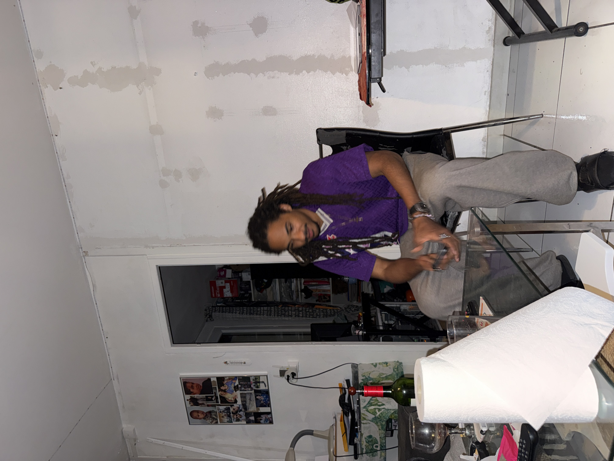 A man with dreadlocks sits in a black chair next to a glass coffee table in a room. He wears a purple shirt and grey sweatpants. The coffee table has a roll of paper towels, a bottle of wine, glasses, and a remote control on it. In the background, there is a doorway leading to another room filled with various items. On the wall near the doorway, there is a collage of photographs. The walls of the room are painted white, with some areas showing signs of repair or patching. The image was taken in Sarcelles, France.