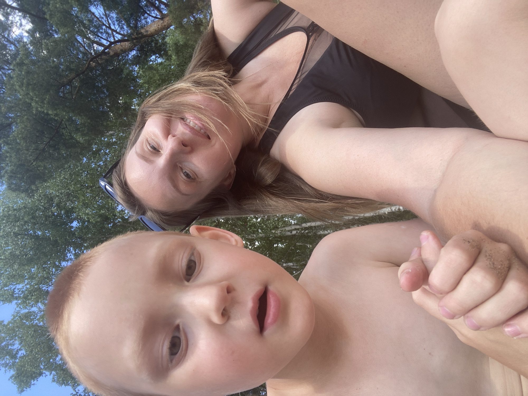 A woman and a young boy are taking a selfie outdoors. The woman is lying down, with her head tilted upwards, and is smiling at the camera. She is wearing a black swimsuit and sunglasses. The boy is in the foreground, also looking at the camera with his mouth slightly open. He is shirtless. They appear to be in a natural setting, with trees and a clear blue sky visible in the background. The lighting suggests it is daytime, and the overall mood is casual and happy. There is no visible text in the image.