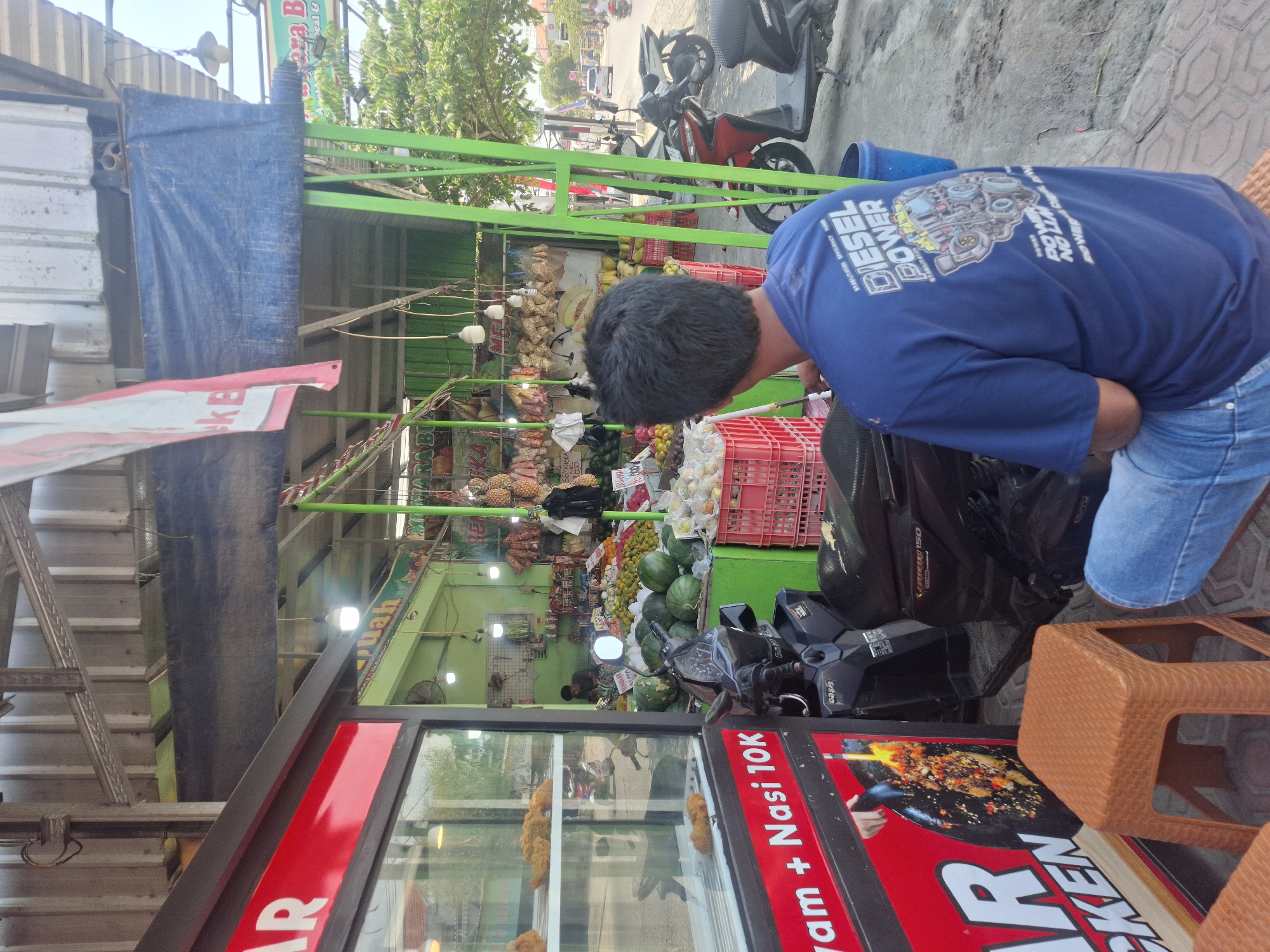 The image depicts a street scene in Sumberwudi, Indonesia. In the foreground, a person wearing a blue t-shirt with a "DIESEL POWER" graphic and jeans is seated on a woven plastic stool. To the left, a food stall is visible, with a red front panel displaying text that includes "ayam + Nasi 10K". Inside the stall's display window, fried chicken pieces are arranged on shelves. A scooter is parked in front of the food stall.
The background reveals a fruit and vegetable market stall with green support poles. A variety of fruits, including watermelons, pineapples, and oranges, are displayed in red crates and on tables. Several motorcycles are parked in the vicinity of the market. Beyond the market, a street with moving vehicles and trees is visible under a clear sky. The overall environment appears to be a typical outdoor market in a town or city.