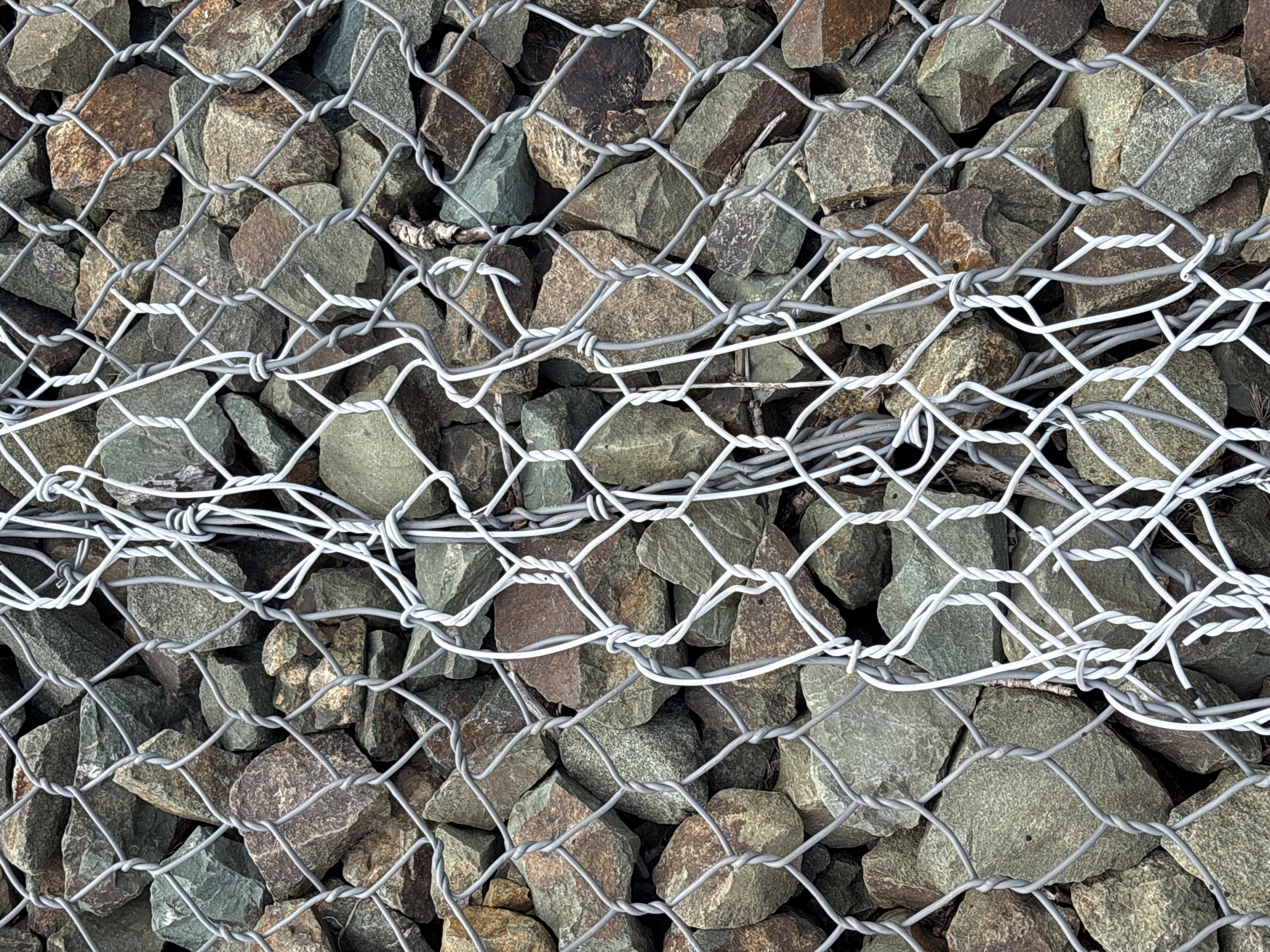 This is a close-up, eye-level, medium shot of a gabion wall filled with rocks, seen through a chain-link fence. The rocks are a mix of earthy tones, including green, brown, and gray. They vary in size and shape, with some being rough and angular, while others are more rounded. The chain-link fence is made of metal wires that are twisted together to form a diamond pattern. Some sections of the fence appear to be reinforced with thicker, white wires. The fence is positioned in front of the gabion wall, obscuring some of the rocks. The overall impression is one of texture and containment. The image does not contain any people or visible signs of specific human activity. The natural elements of the rocks and the manufactured elements of the fence create a visually interesting contrast. It is impossible to determine the specific location within Orenburg, Russia from the provided image.