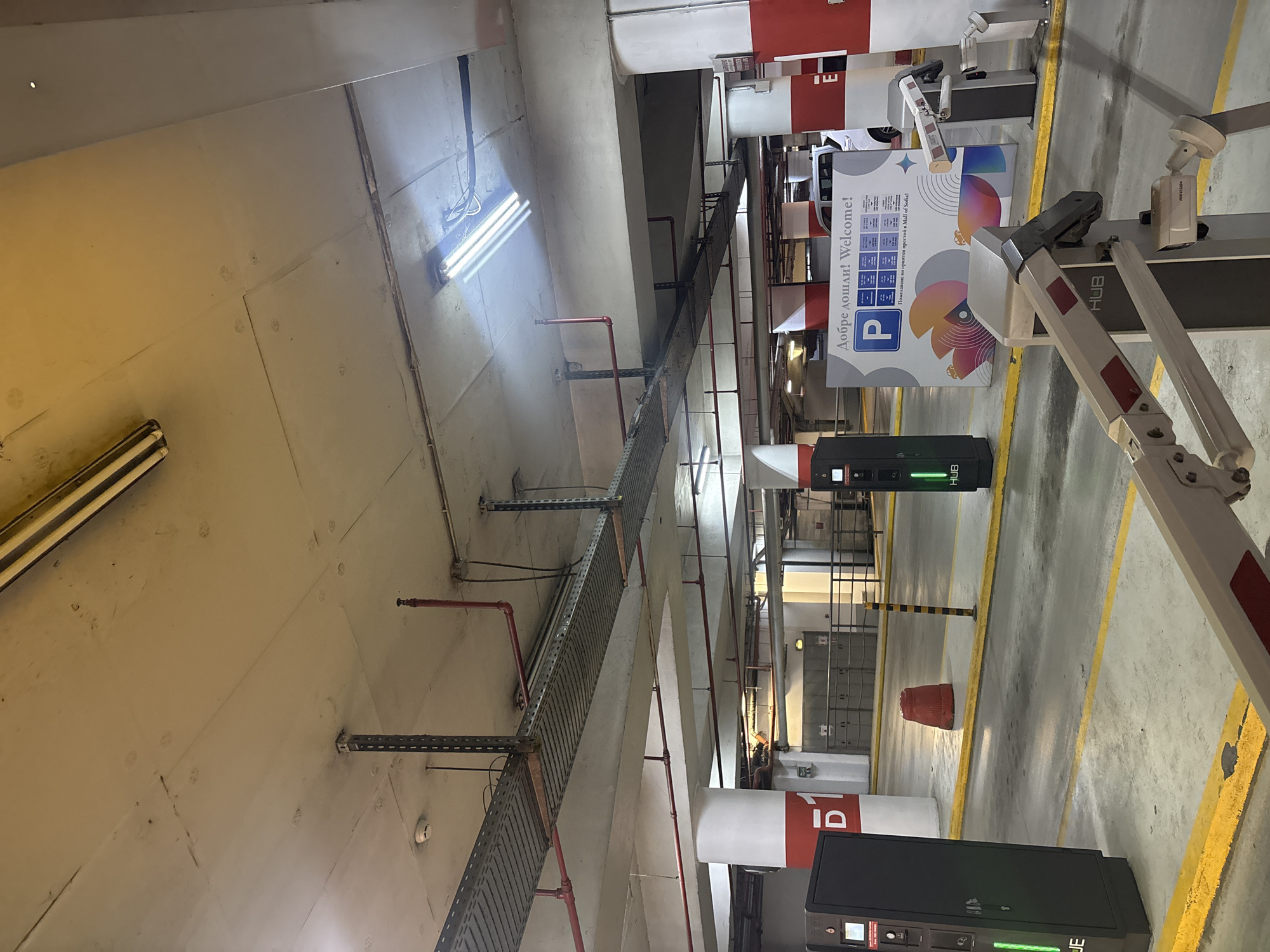 The image shows a parking garage with fluorescent lighting and concrete pillars. On the left, there is a black payment kiosk with a green light indicator. In the middle ground, a parking boom barrier is partially visible in the foreground. Further back, a large sign with the text "ДобPе дошли! Welcome!" and a parking symbol is displayed. Behind the sign, a white SUV is parked. To the right, another payment kiosk and a security camera are visible. The environment appears to be well-lit and functional, indicating a typical indoor parking facility.