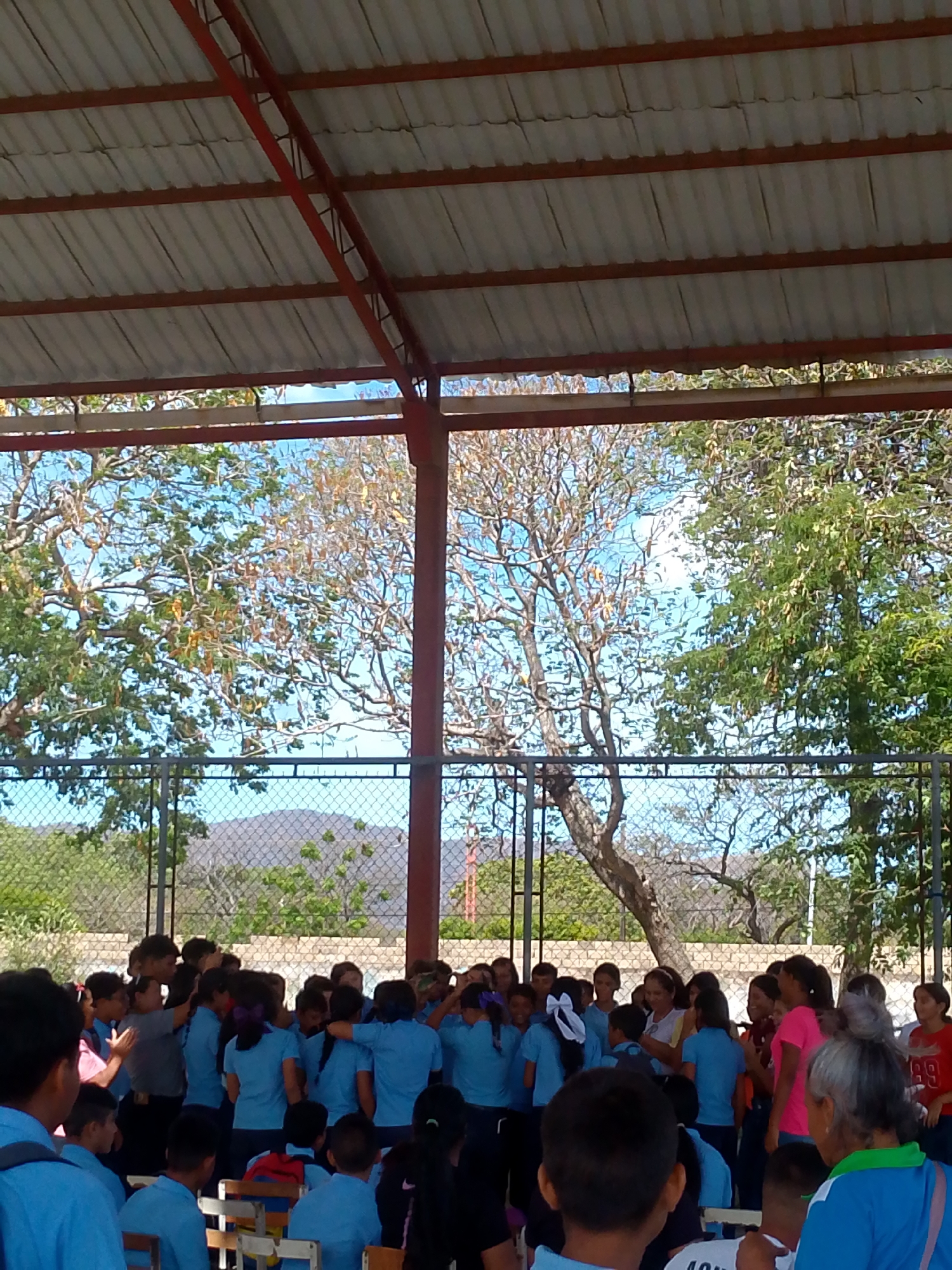 The image shows a group of people, mostly children dressed in blue shirts, gathered under a large, open-sided structure with a metal roof supported by red metal beams. The location is likely Juan Griego, Venezuela, given the presence of what appears to be local school children.
Behind the group, there's a fence separating the area from trees and a mountain range in the distance. The sky is visible through the trees. Some adults are also present, including one wearing a pink shirt and another with grey hair wearing a green-trimmed shirt. The children are clustered together, and it looks like they might be interacting with the adults. Many of the children are seated on wooden chairs.