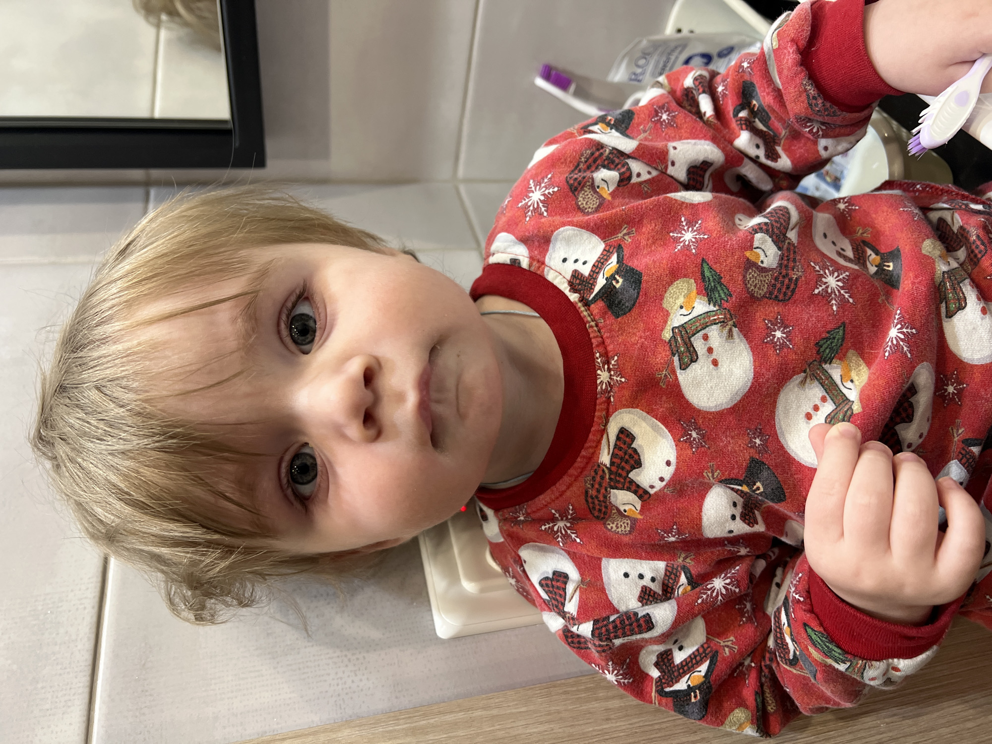A young child with fair skin and blonde hair, wearing a red long-sleeved shirt with a snowman pattern, is looking at the camera. The child's face is partially visible, showing their eyes, nose, and mouth. A white toothbrush is visible in the child's hand. The background includes a white tiled wall, a mirror, and a wooden countertop. The location is not Noviy, Russia.