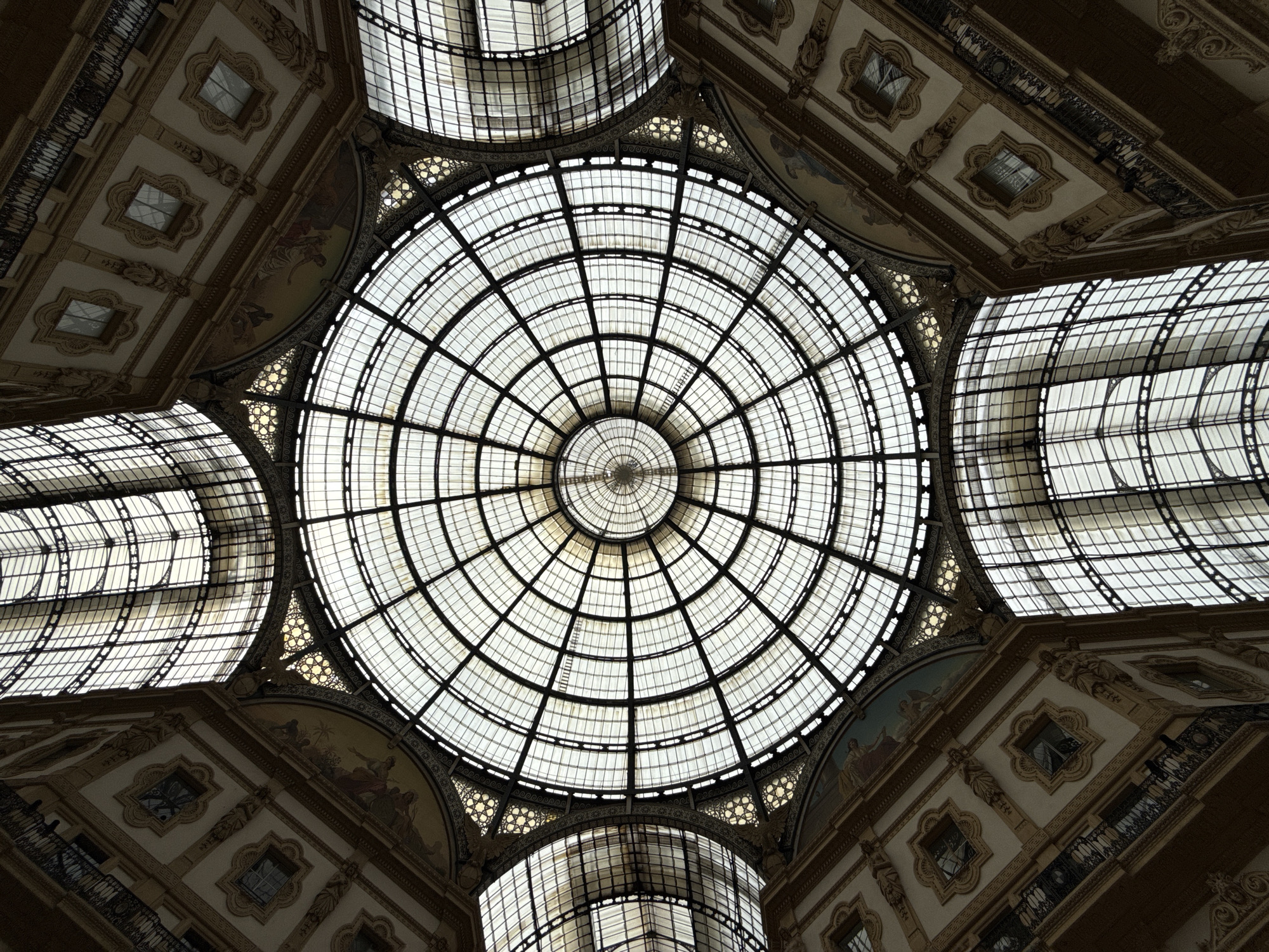 The image captures an upward view of the glass dome ceiling within the Galleria Vittorio Emanuele II, a historic shopping arcade located in Milan, Italy. The dome is composed of a circular grid of glass panes and metal supports, with a central oculus at its peak. The architecture surrounding the dome features ornate detailing, including painted murals, decorative moldings, and small windows or openings. The perspective is from below, looking directly up at the intricate structure.