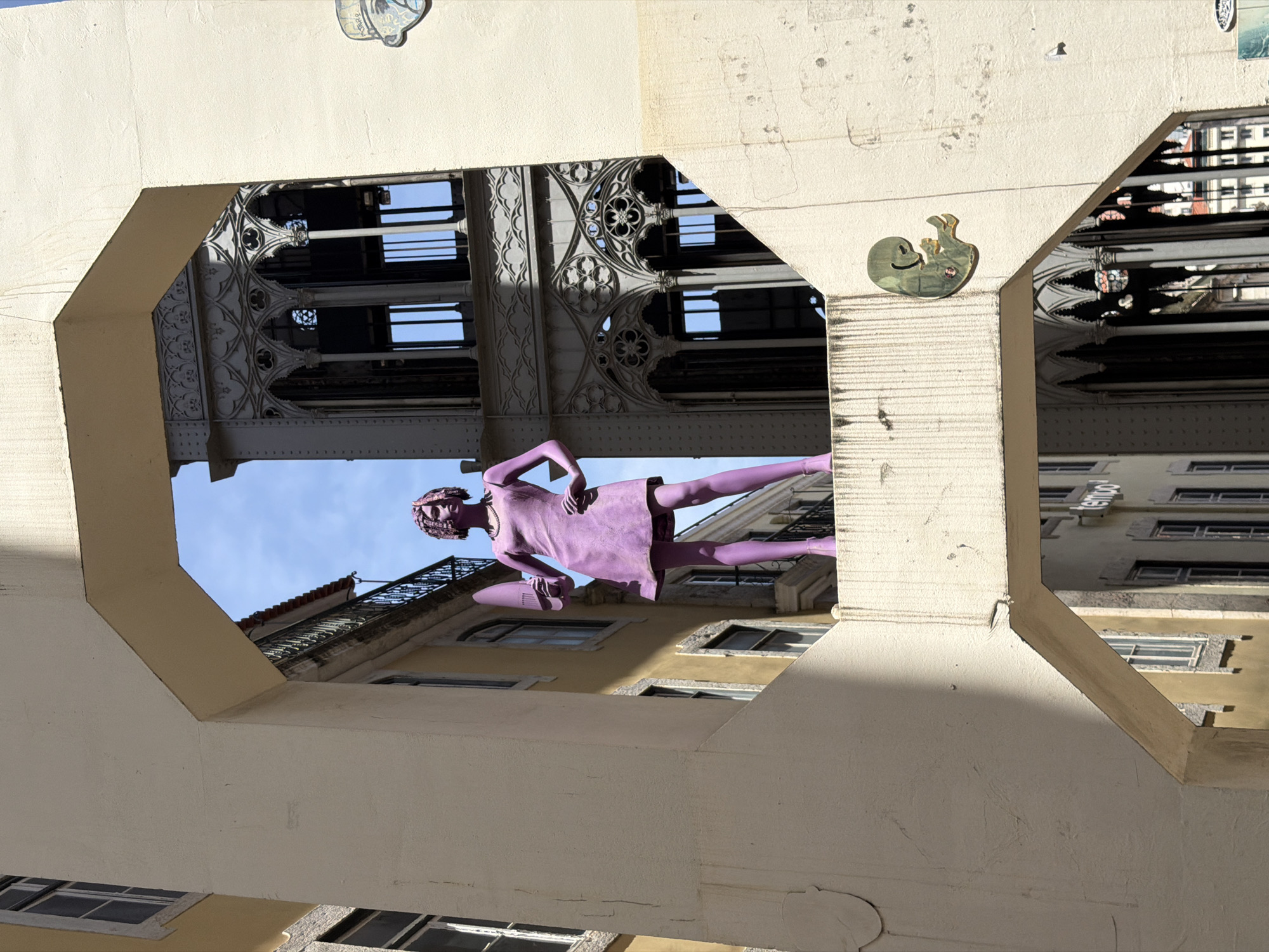 A bright, sunny day in Lisbon, Portugal, offers a view through an angular, cream-colored concrete archway. The arch frames a vivid purple statue of a woman standing on a ledge. She wears a short, sleeveless dress and heels, with her left hand on her hip and her right hand holding a large, dark object, possibly a hair dryer. Her hair is styled in a textured, bob cut. Behind her, an ornate, dark metal structure with Gothic-inspired architectural details rises. In the background, the blue sky is visible, along with the facades of yellow and cream-colored buildings, some with balconies and windows. The concrete structure also features a small, golden emblem of a fetal silhouette attached to its surface. The overall scene suggests an urban art installation integrated into the city's architecture.