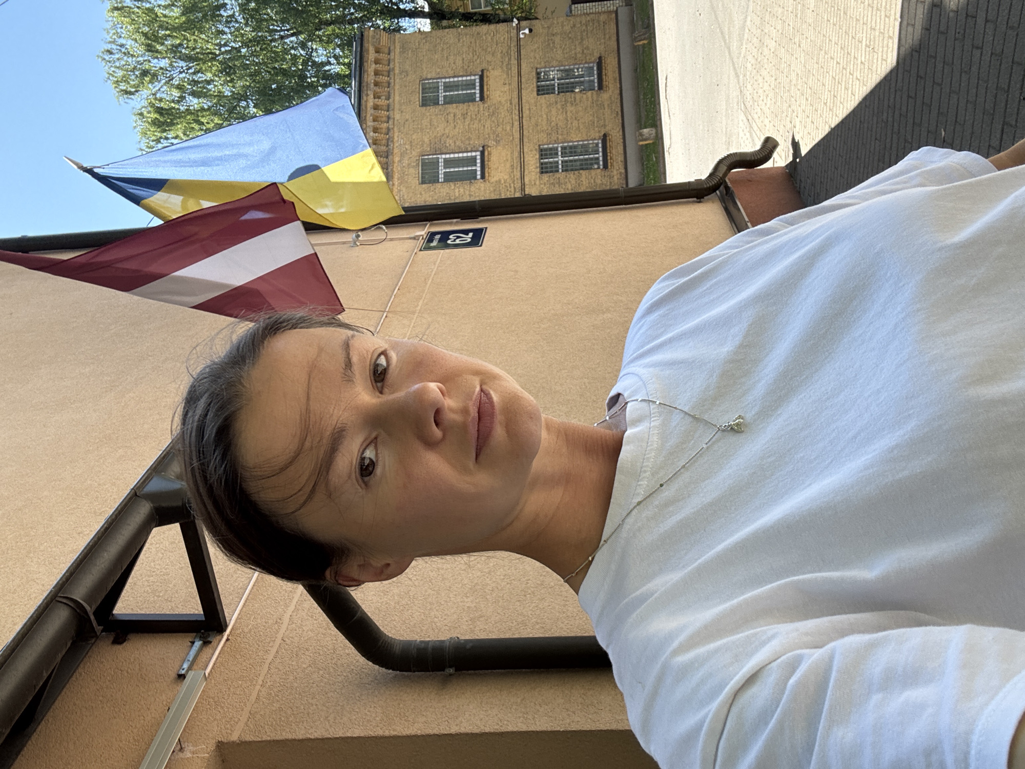 A woman with dark hair pulled back and wearing a white t-shirt and a silver necklace stands outdoors. She is taking a selfie. Behind her is a beige building, with a downspout and some flags attached to the wall. The flags are the national flags of Latvia and Ukraine. A yellow brick building with windows is visible behind them, and a tree with green leaves is to the right. The ground is paved with gray bricks. A blue sign with the number 62 is on the wall. The lighting suggests it is daytime with strong sunlight, casting shadows.