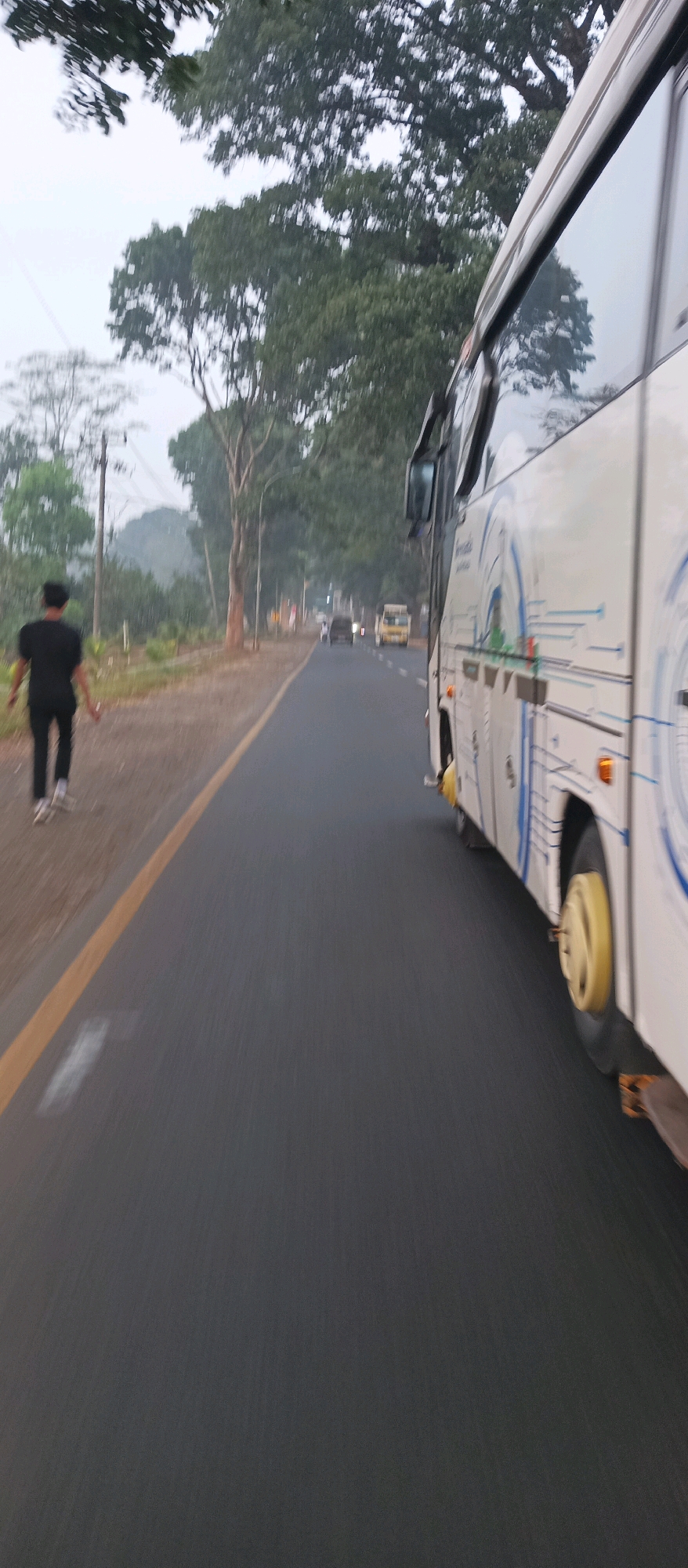 A person in a black shirt and pants walks along a paved road in Jalancagak, Indonesia, beside a white bus. The road is asphalt with a yellow line marking the edge. Trees and foliage line the left side of the road, with a utility pole visible in the distance. Other vehicles, including a yellow bus and a dark car, are seen further down the road. The sky is overcast, suggesting it is either early morning or late afternoon. The overall scene depicts a typical roadside view on a cloudy day in a semi-rural Indonesian setting.