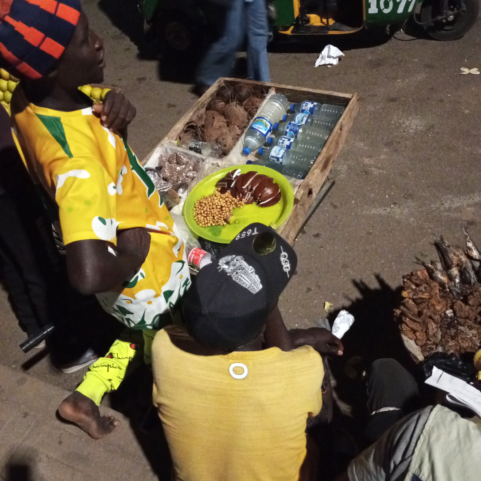 A street scene in the city of Unknown, Unknown, shows at least three individuals near a wooden cart stocked with various goods for sale. An individual in a blue and red striped hat and a yellow shirt with green and white patterns stands to the left of the cart. The cart contains coconuts, bottled water, a green plate with what appears to be liver and beans, and other packaged snacks. To the right of the cart, an individual wearing a black cap with a design and a yellow shirt is seated. Next to them is a pile of dried fish. Another individual, partially visible, is seated near the fish. The ground is paved, and a green vehicle with the number 1077 on it is visible in the background.