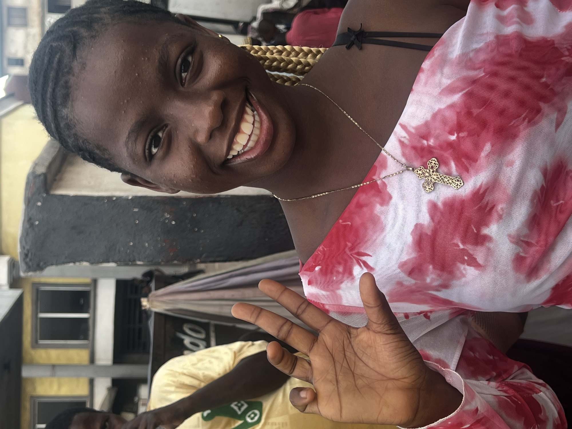 A young Black woman with dark skin and braided hair smiles widely, her mouth open to reveal white teeth. She wears a light-colored, loosely woven top with a pink tie-dye pattern and a delicate gold necklace with a cross pendant. Her left hand is raised, palm facing the camera, with fingers spread apart. She is wearing a small gold stud earring in her visible ear. In the background, a man in a yellow shirt stands partially visible to her left. Behind them, a building with yellow siding and dark-rimmed windows is visible, as is a dark, textured wall and what appears to be draped fabric. The scene appears to be outdoors in a populated area, possibly a market or street in Nkpor, Nigeria.