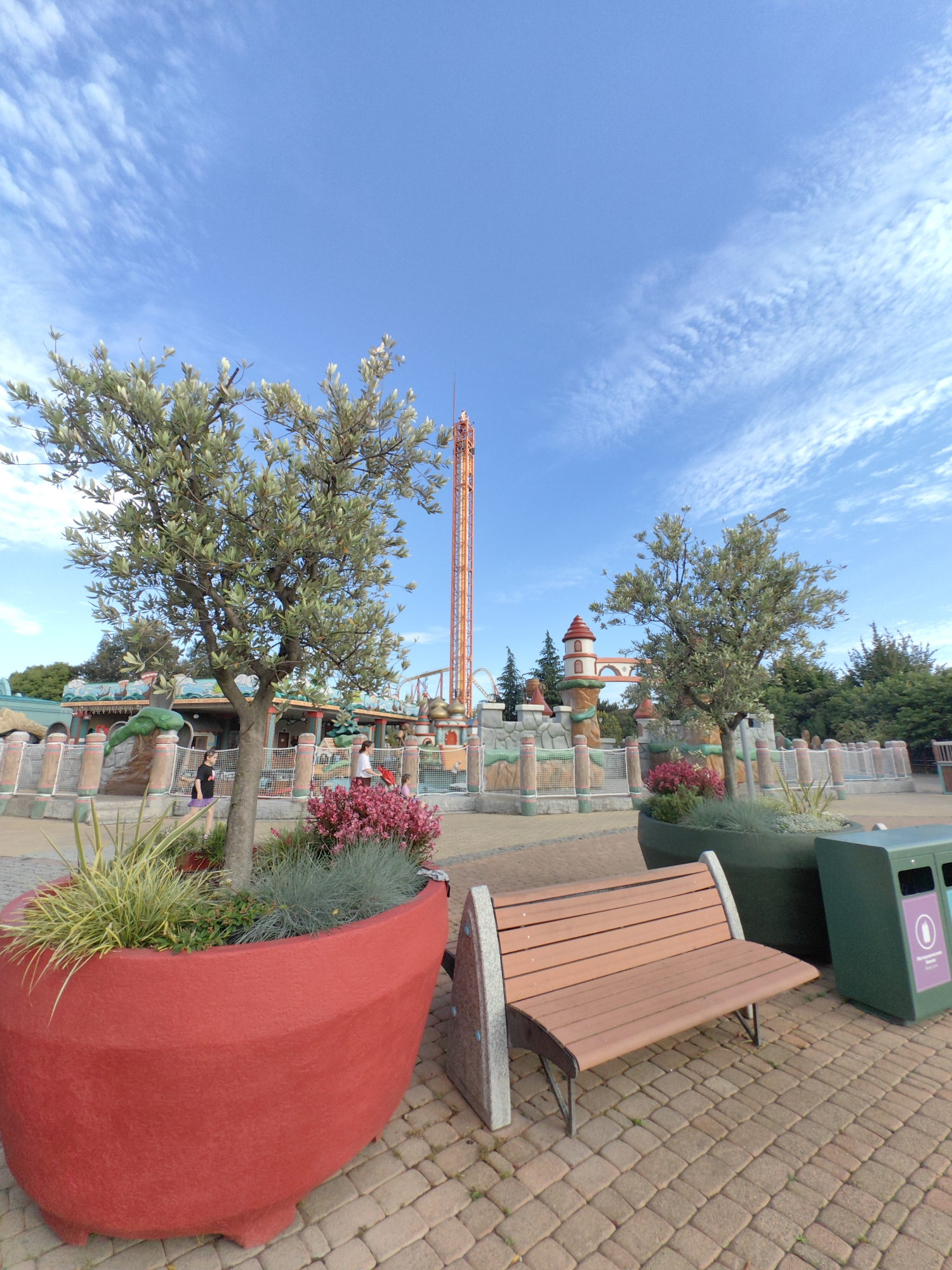 An outdoor, eye-level, wide shot captures an amusement park scene on a clear, blue-sky day with sparse, wispy clouds. In the foreground, a large, bright red planter overflows with lush green plants and small pink flowers. To the right of the planter, a wooden bench with a metallic frame sits on a cobblestone path. Behind the bench is a green trash receptacle. Further back, a mature tree with silvery-green leaves stands tall. Beyond the tree, an amusement park attraction, resembling a drop tower, with an orange structure and a visible ride mechanism, dominates the background. The attraction is surrounded by colorful, whimsical structures, reminiscent of a fairytale castle, with turrets, arches, and decorative fencing. Several people are visible in the mid-ground, walking and interacting within the park. The environment appears to be a well-maintained, vibrant amusement park.