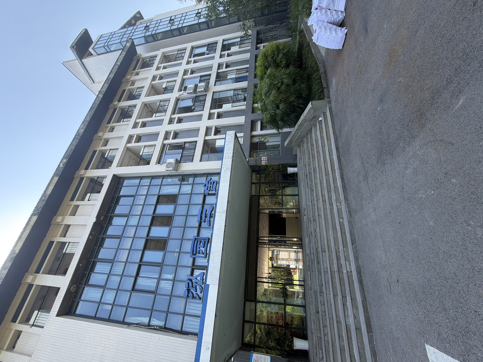 The image depicts the exterior of a multi-story building, likely a dormitory or administrative building, in Jiangjin, China. The building has a modern architectural style with a facade featuring large glass windows and white tiled sections. Blue Chinese characters, which translate to "Hongyuan Hall," are prominently displayed above the entrance. A wide set of concrete stairs leads up to the main entrance, which consists of glass doors. The surrounding environment shows a paved area in front of the stairs, some greenery, and a stack of white bags on the right side of the image. The sky is clear and blue, suggesting daytime. There are no people visible in the image. The scene appears to be a calm and ordinary day at this location in Jiangjin.
