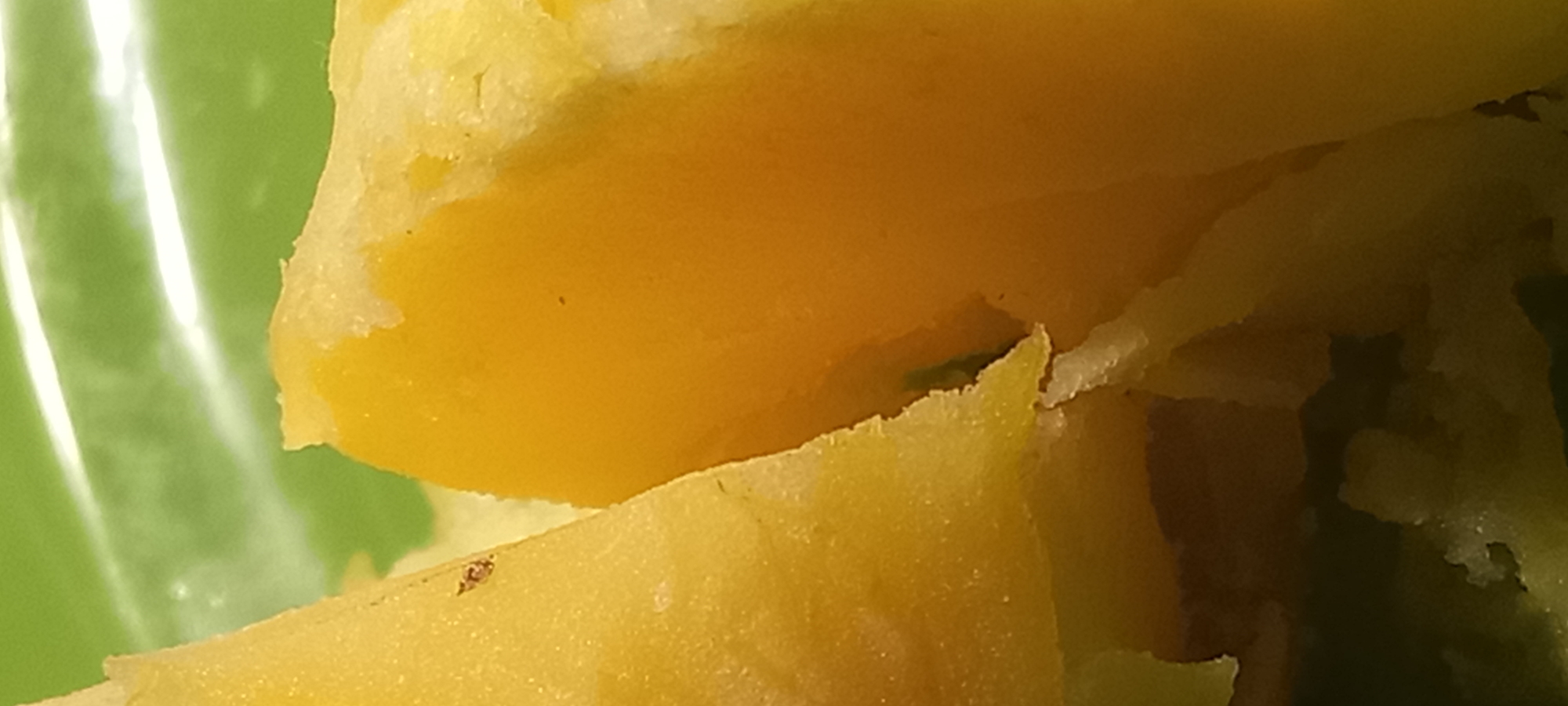 The image shows a close-up, macro shot of food, specifically pieces of cooked potato. The potatoes are cut into irregular chunks, revealing a fluffy, pale yellow interior with slightly browned edges. One piece on the left is larger and shows a slightly rough, textured surface with a few small dark specks. The potato on the right is a smooth, solid block of yellow.
The potatoes are presented against a bright, lime green background which appears to be a surface, possibly a plate or tablecloth, with a glossy finish, as indicated by the bright white highlights reflecting light. There are no discernible environmental details that would allow for a specific location to be identified within Kryvyi Rih, Ukraine, or any other city. The focus is solely on the food items. There are no people or actions depicted.