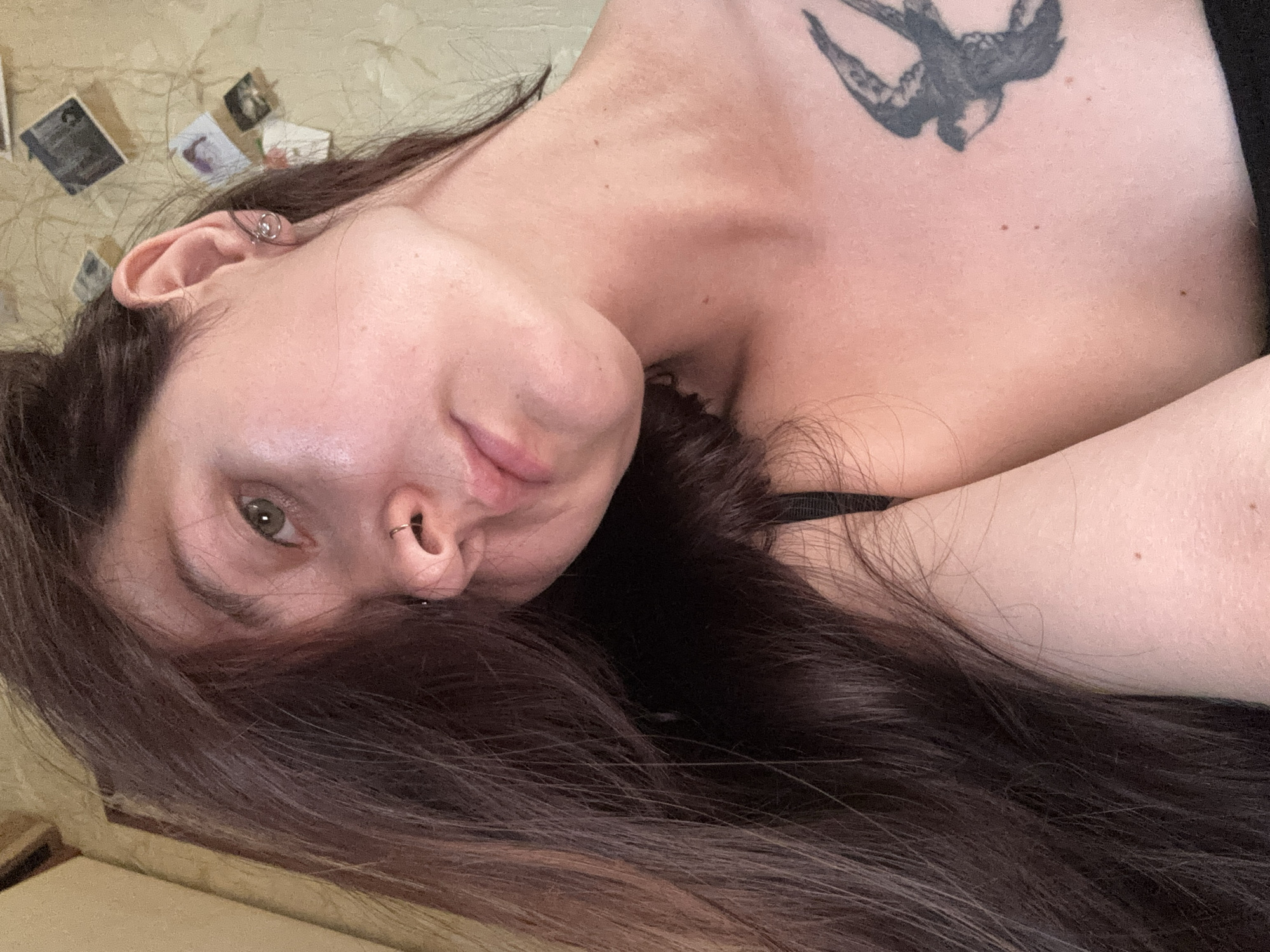 The image is a close-up, angled selfie of a young woman with long, dark brown hair covering part of her face. She has fair skin, green eyes, and a nose ring. She is wearing a black bra strap and has a tattoo of a bird on her chest. The background appears to be an interior wall with several small framed pictures hanging on it. There is no indication of the city of Naberezhnye Chelny, Russia, in the image.