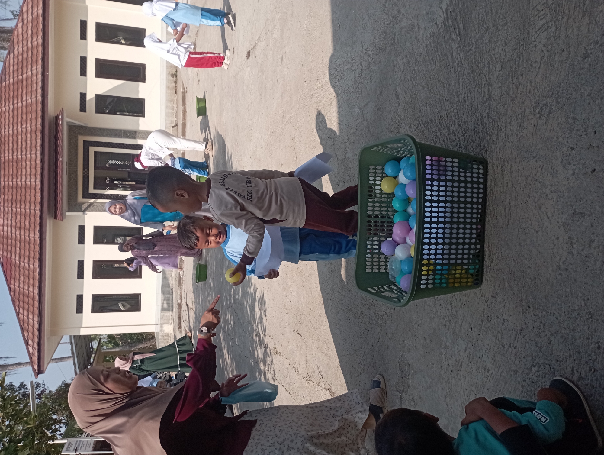 A group of children and adults are gathered outdoors in what appears to be a schoolyard or event space. A woman in a brown hijab and maroon top is pointing at a young boy in a blue shirt who is holding a yellow ball. To the boy's right, another boy in a light brown shirt is holding a white paper. In the foreground, a green basket is filled with colorful plastic balls. Several other children and adults are visible in the background, some wearing white uniforms and headscarves. The scene is set against a backdrop of a building with a brown tiled roof and a clear blue sky. The ground is paved with concrete. The image does not provide specific information about its location in Garut, Indonesia, but the context suggests an educational or community event.