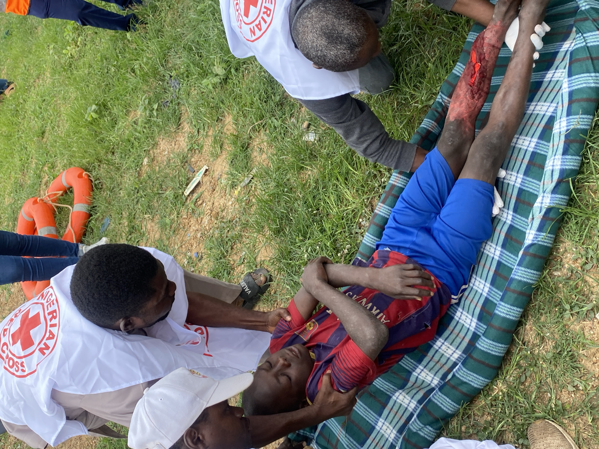 In Kawo, Nigeria, a young boy lies on his back on a green and blue checkered blanket spread on the ground. He is wearing a red and blue striped shirt and blue shorts, with dirt or dust covering his arms and torso. His left arm is bent across his chest, and his right arm is extended. A significant laceration is visible on his right lower leg, exposing muscle tissue and what appears to be a piece of red material.
Two individuals wearing white vests with the Red Cross emblem are attending to the boy. One person, wearing a white cap, is kneeling beside the boy's head and shoulders, with their hands gently placed on him. Another individual, also wearing a Red Cross vest, is kneeling at the boy's feet, wearing white gloves and tending to his injured leg. In the background, two orange lifebuoys lie on the grass. The scene is outdoors, with green grass and some brown earth visible. A person in blue jeans and a white shirt is partially visible in the upper background, standing near the lifebuoys.