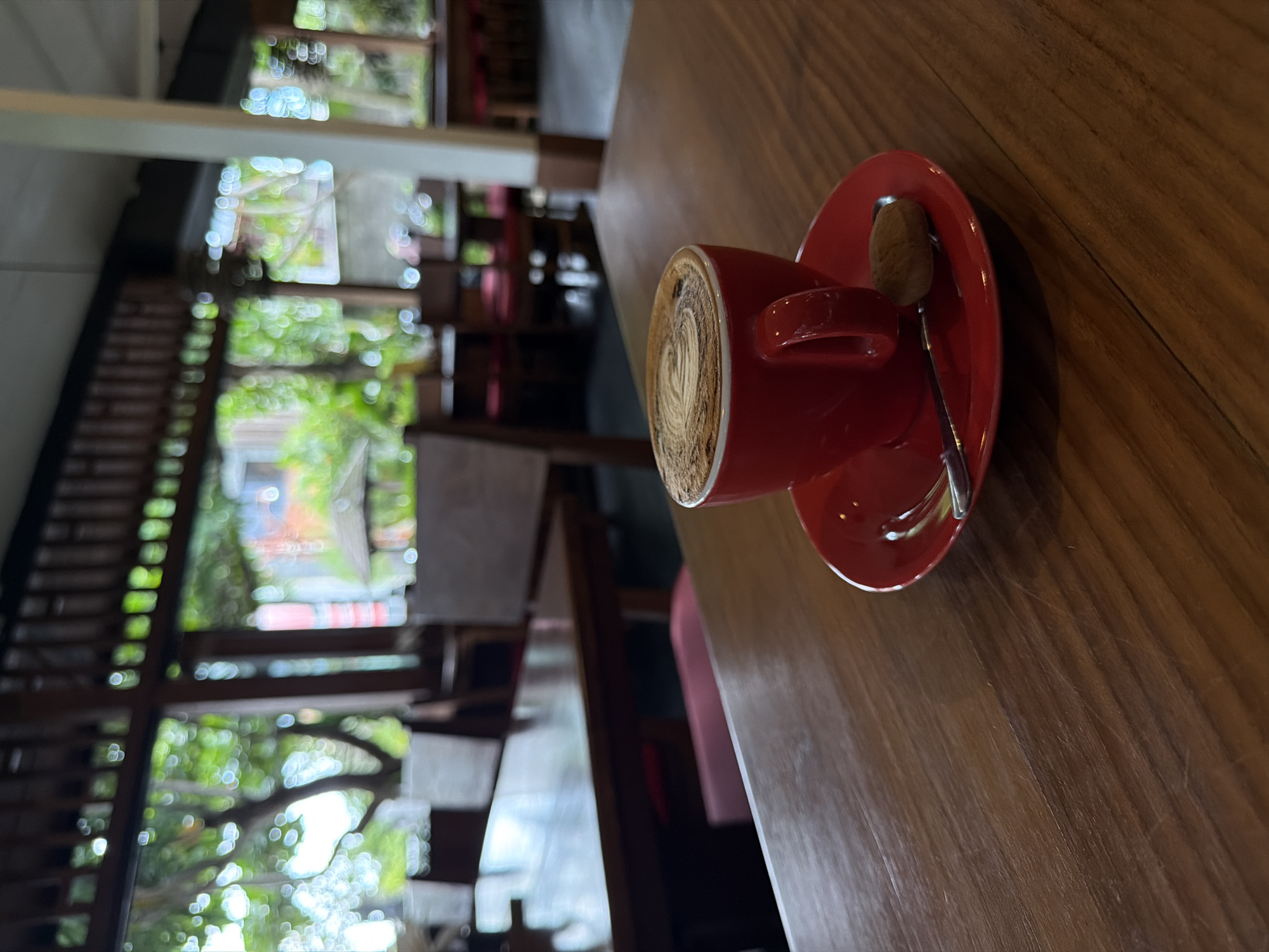 A red cup and saucer with a spoon rest on a wooden table. The cup contains coffee with latte art. The background shows a blurred outdoor area with trees and buildings, indicative of an open-air café or restaurant setting. The location is in North Kuta, Indonesia.