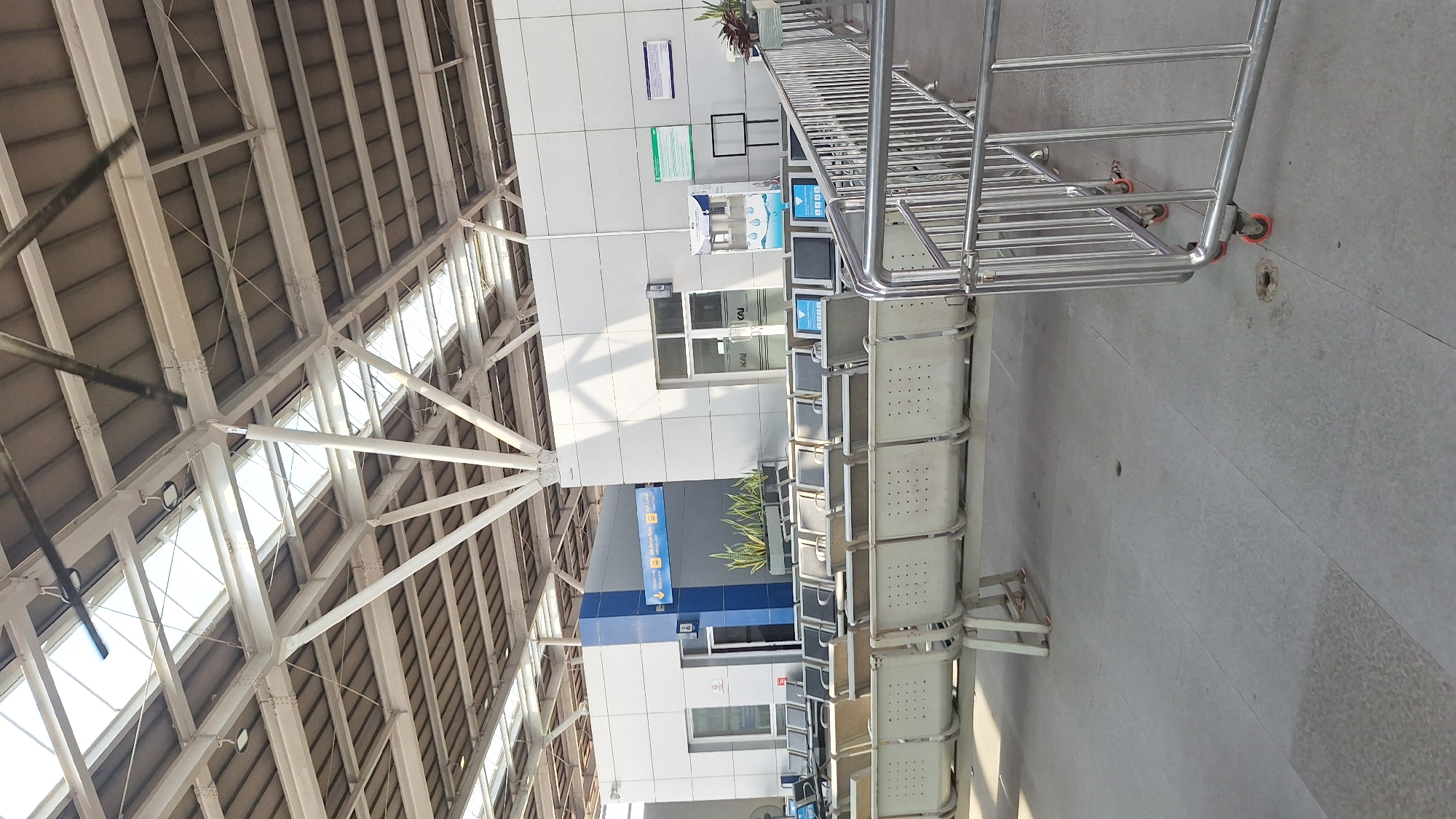The image shows the interior of a train station waiting area in Cikarang, Indonesia. The ceiling is high, with a complex network of white metal beams and translucent panels providing natural light. A row of light gray plastic chairs lines the right side of the frame, leading towards a set of metal barriers on wheels. A wall runs along the back, with signage, windows, and doors. A potted plant sits atop a partition near the front. The overall scene appears clean and well-maintained, with a predominantly gray and white color scheme.