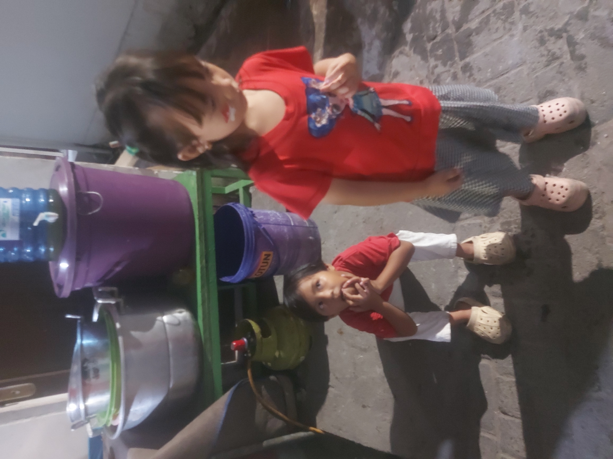 The image depicts two young children outdoors, possibly in the city of Makassar, Indonesia. In the foreground, a girl in a red t-shirt with a cartoon character print and a checkered skirt stands facing right. Her face is slightly blurred, but a smudge of white and red paint is visible on her cheek. She wears pink clogs and has a green hair accessory. Beside her, a younger child in a red t-shirt and white pants kneels on the ground, looking upwards with their hands near their mouth. This child also wears beige clogs.
In the background, there is a green shelf holding several metal bowls and a purple bucket with the word "OTUN" visible. A large blue water jug sits atop a purple container behind the shelf. To the left of the shelf, a green gas cylinder with a yellow hose is visible. The ground appears to be paved with concrete tiles, and shadows are cast by the children and objects. The lighting suggests it is either early morning or late evening.