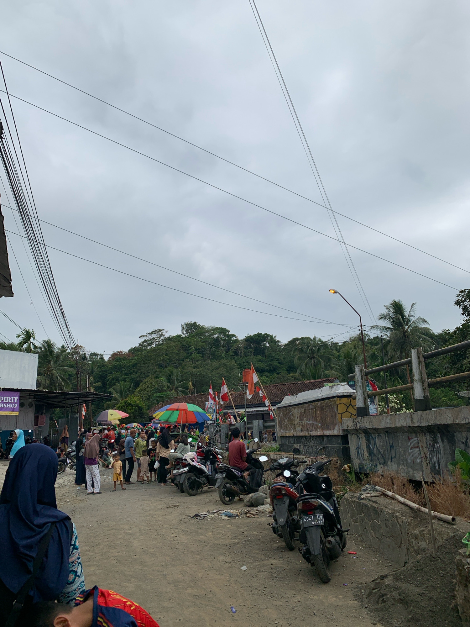 The image depicts a street scene in Karangmoncol, Indonesia, on a cloudy day. The foreground shows a dirt road with several parked motorcycles. In the middle ground, a crowd of people are gathered, some standing and talking, others walking. Colorful umbrellas are set up, likely for vendors or shade. Indonesian flags are visible, suggesting a national celebration or event. The background consists of lush green hills and trees, with palm trees also present. A concrete bridge with a railing spans across a section of the scene. Electrical wires crisscross the sky, and a street light is illuminated. The overall impression is of a lively, casual gathering in a semi-rural or suburban area.