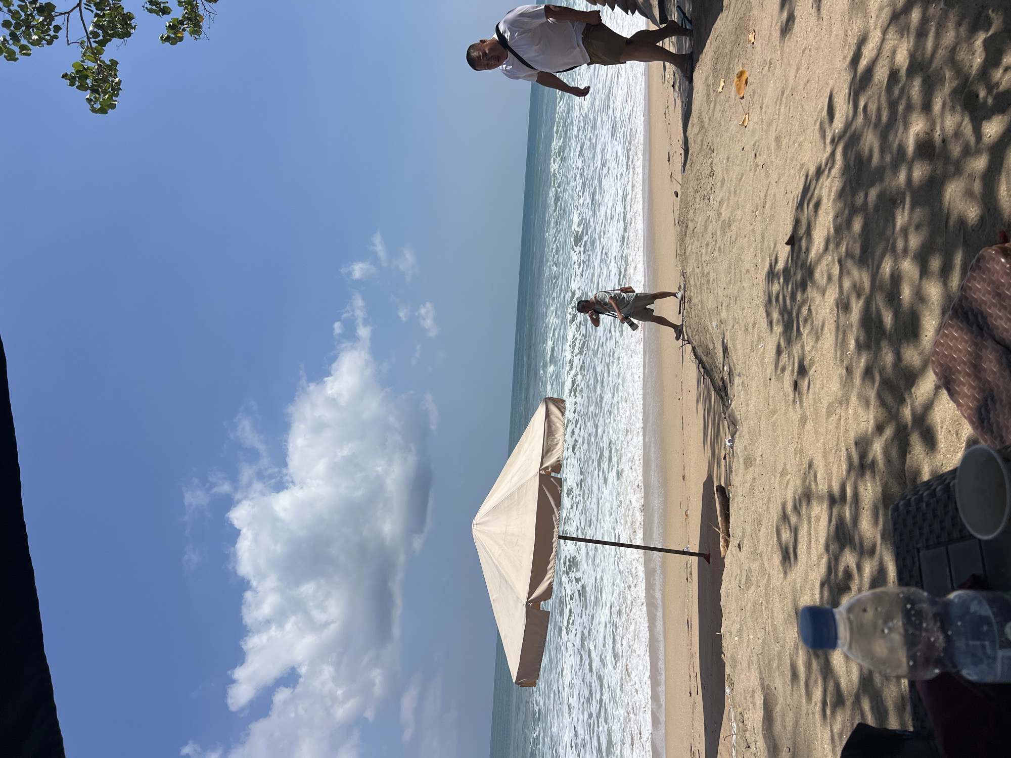 An eye-level, medium shot captures a beach scene in Kuta, Indonesia, on a sunny day. The sandy shore is the foreground, leading to the ocean with its foamy waves. A large, light-colored beach umbrella stands prominently on the sand. Two men are visible walking along the beach, with one in the foreground on the right, wearing a white shirt and shorts, and another in the mid-ground on the left, carrying a camera. The sky is blue with scattered clouds, and tree branches with green leaves are visible at the top right. In the lower foreground, a plastic water bottle and a cup rest on a woven surface, suggesting a seated observer.