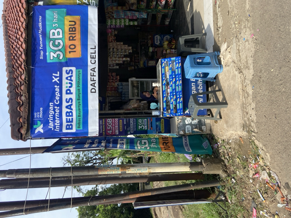 The image shows a small storefront in Jatiluhur, Indonesia (latitude: -6.571, longitude: 107.44). The storefront is painted in a light blue color and has a large sign above it advertising "Jaringan Internet Cepat XL Bebas Puas" and "3GB 3 hari 10 Ribu" with the words "DAFFA CELL" at the bottom. The storefront is selling cellular phone data packages and other items. There is a woman sitting on a blue and white stool in front of the storefront. She is wearing a light blue headscarf and a white shirt. The storefront has a glass display case with various items for sale. There are two stools sitting on the sidewalk in front of the storefront. There is a sign hanging from the ceiling that says "we are more". The ground is covered with dirt and debris, including plastic bottles, bags, and other trash. There is a dirt road and some trees visible behind the storefront.