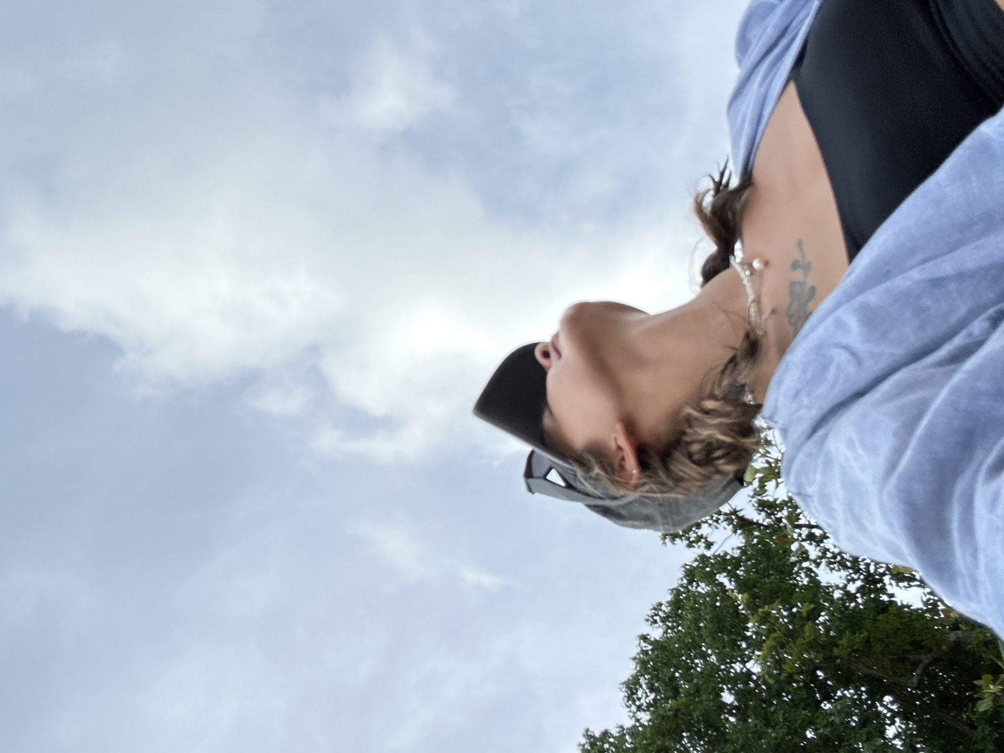 A low-angle shot captures a person from the shoulders up, looking skyward against a cloudy sky. The person is wearing a black tube top, a light blue open cardigan, and a grey baseball cap. A pearl necklace is visible around their neck, and a tattoo is on their collarbone. To the left, the branches of a dense green tree are visible. The scene appears to be outdoors, possibly in the region of Phru Nai, Thailand, given the tropical foliage.