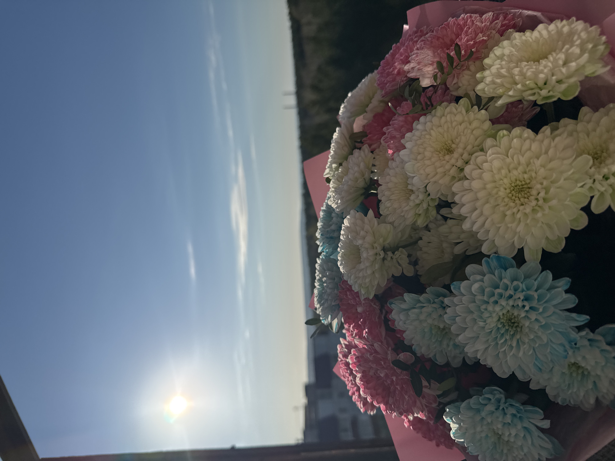 The image shows a bouquet of flowers in shades of white, pink, and blue, with pink wrapping paper. The flowers are held against a backdrop of a clear, blue sky with the sun visible in the lower left corner. In the distance, a faint horizon line suggests a landscape. Based on visual clues, the location is Chaikovsky, Russia. The composition is slightly tilted, with the bouquet positioned to the right of the frame. The overall scene appears to be taken outdoors, possibly on a balcony.