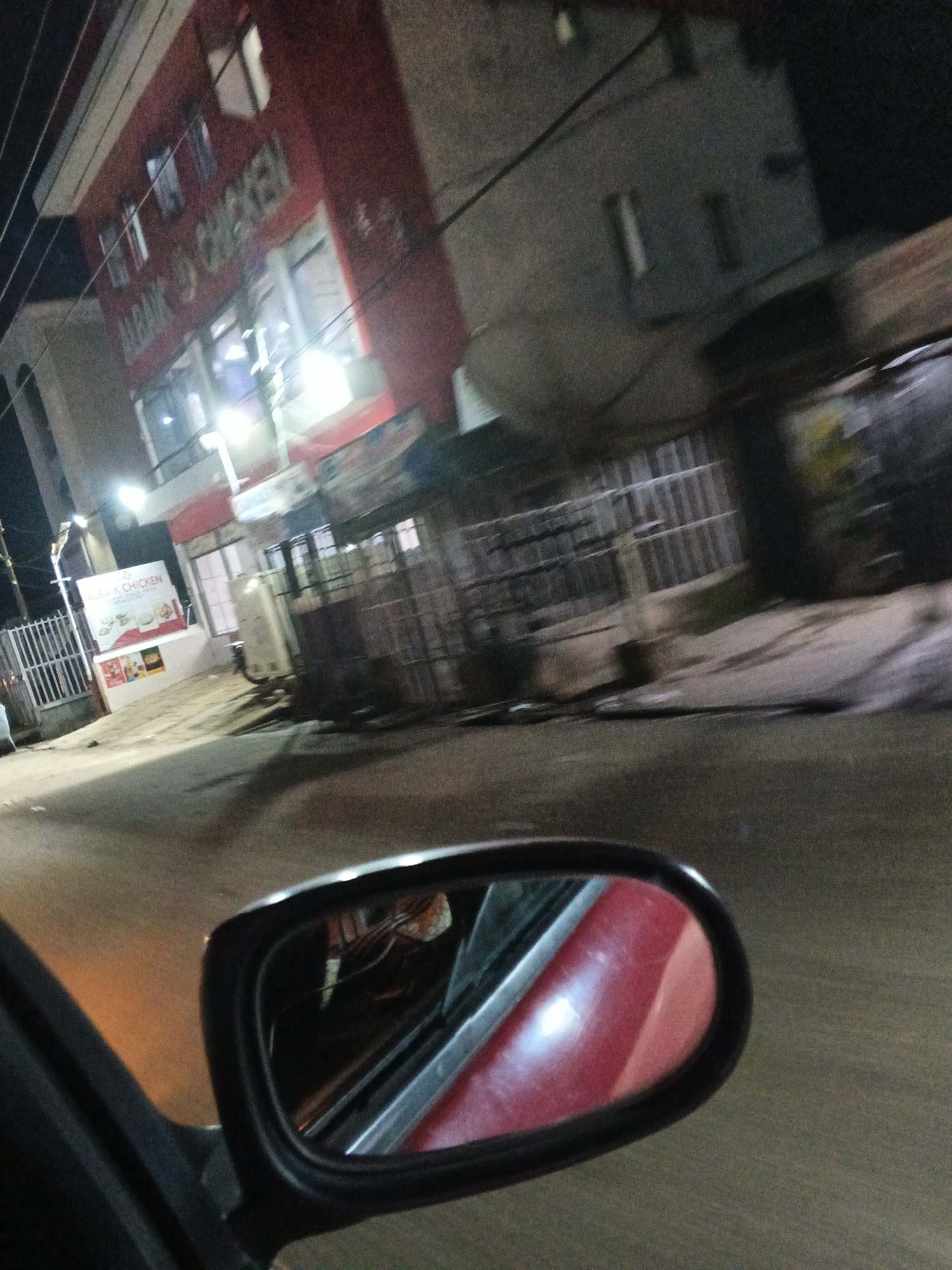 An outdoor, eye-level, medium shot captures a street scene at night, likely in Jos, Nigeria, given the presence of "ALBAIK CHICKEN" signage. The image is taken from within a vehicle, as evidenced by the rearview mirror reflecting the interior of the car, featuring a red seat. The foreground is dominated by the car's side door and the rearview mirror.
The background shows a multi-story building with red and grey facades. The building's signage, "ALBAIK CHICKEN," is prominently displayed in the upper left quadrant, illuminated by lights. A smaller sign below it, also for "ALBAIK CHICKEN," includes images of food. Adjacent to the building is a white fence. The street in front of the building appears wet, reflecting the ambient light. The overall impression is one of a busy commercial area at night, possibly a fast-food establishment.