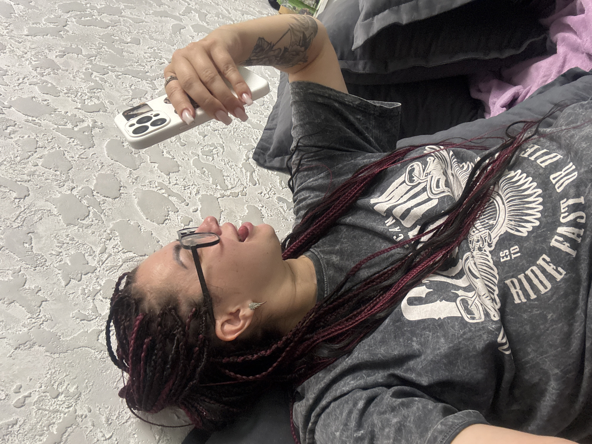 In an indoor setting with a textured light grey wall, a woman with dark braided hair, some strands of which are burgundy, is visible. She is wearing black-rimmed glasses and a dark grey t-shirt with a white graphic print that reads "RIDE FAST OR DIE". Her tongue is sticking out playfully as she holds up a white smartphone in her right hand, seemingly taking a selfie. She is also wearing a silver earring in her left ear and a ring on her right ring finger. The woman is leaning against dark grey pillows, and a purple blanket is partially visible. There is a tattoo on her right arm. No specific location within Tolyatti, Russia, can be identified from the provided image.