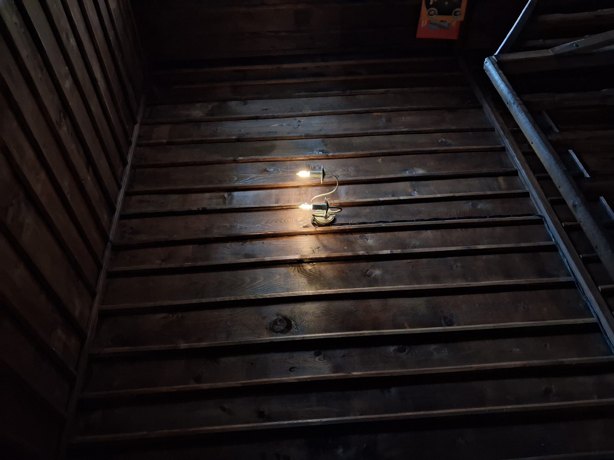 The image depicts the interior of a wooden structure, likely a building, in Katunishte, Bulgaria. The walls and ceiling are constructed from horizontal wooden planks, creating a dark, rustic environment. A two-bulb wall-mounted light fixture illuminates a section of the wall, casting a warm glow. To the right, a wooden ladder or support structure is visible against the wall. The lighting is dim, and the overall scene is characterized by deep shadows and the texture of the wood.