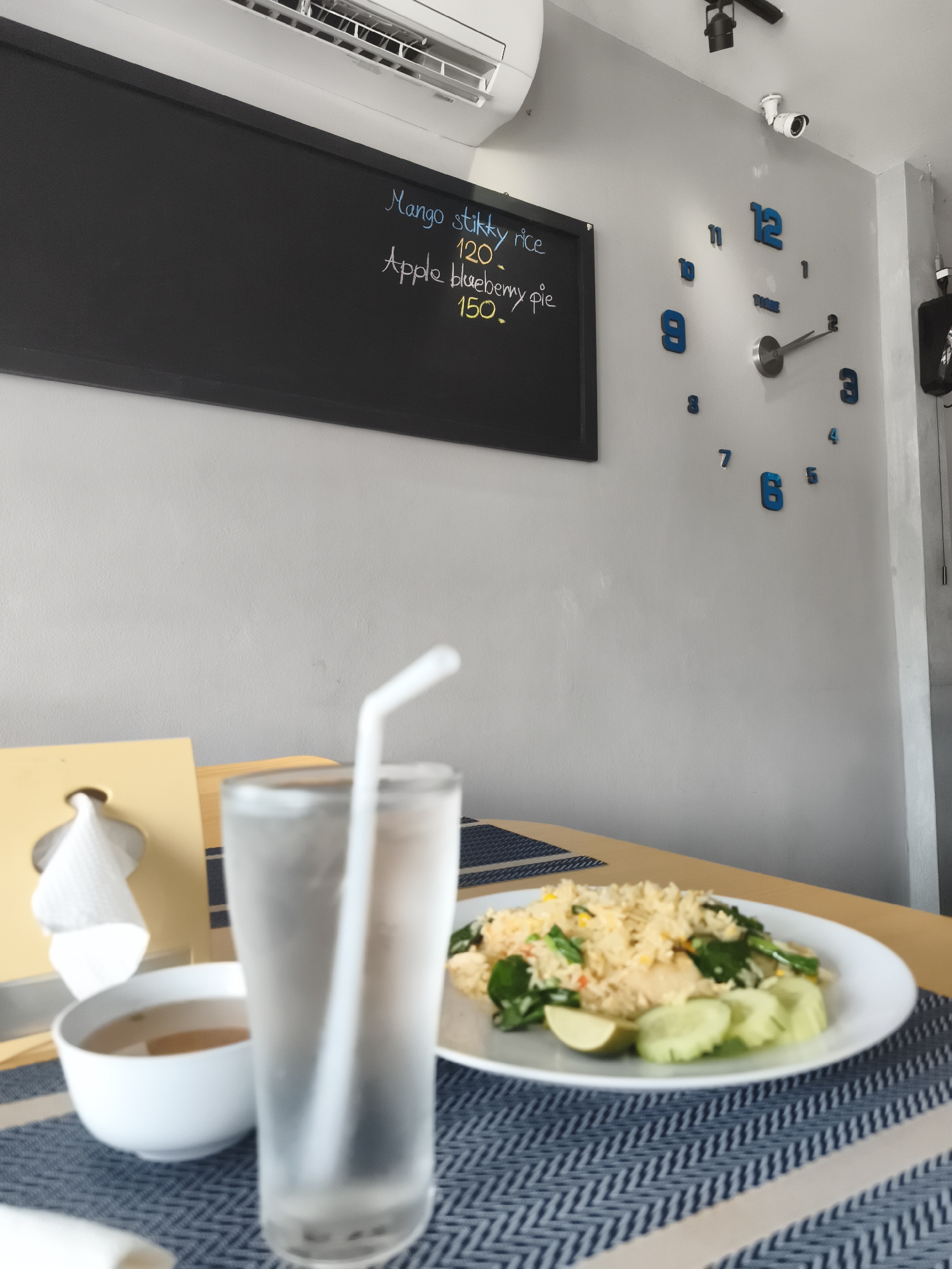 This image depicts an indoor dining scene, likely within a cafe or restaurant in Baan Bang Rak, Thailand. In the foreground, a wooden table is set with a blue and white patterned placemat. A tall glass of water with a white straw is prominently placed, along with a small white bowl, possibly containing soup or sauce. To the right, a white plate holds a portion of fried rice, garnished with green vegetables, cucumber slices, and a lime wedge. A yellow tissue dispenser is also visible on the table.
The background features a light grey wall. Mounted above is a white air conditioner. Below it, a black rectangular blackboard displays a handwritten menu in chalk. The visible text lists "Mango stikky rice 120." (with "sticky" misspelled as "stikky") and "Apple blueberry pie 150.". These items, particularly "Mango sticky rice," are characteristic of Thai cuisine, aligning with the provided location context. To the right of the blackboard, an artistic wall clock with separate blue numeral cutouts, including "TIME" at its center, indicates approximately 2:10 or 2:11. A white security camera and a black spotlight are mounted near the ceiling above the clock. The scene conveys a calm, ready-to-eat atmosphere.