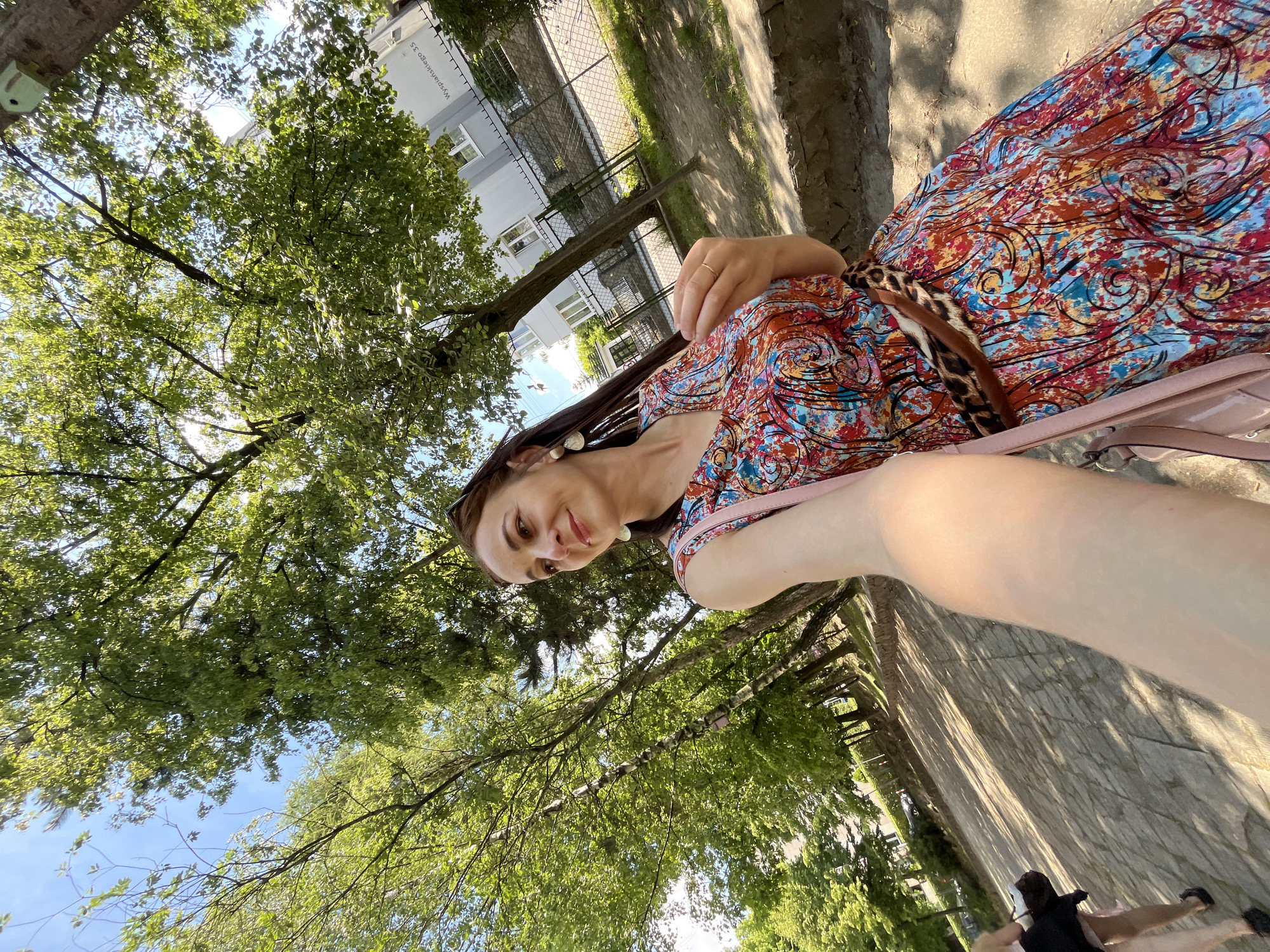 A woman is taking a selfie outdoors in a park or natural setting. She is wearing a brightly colored, patterned dress and has long dark hair. Her right arm is extended, holding the camera. She is smiling and looking towards the camera. The background shows lush green trees with sunlight filtering through the leaves, and a blue sky above. To the right of the woman, a paved path and a stone wall are visible. In the distance, a building with windows and a fence can be seen. The lighting suggests it is daytime, likely sunny. The overall mood is cheerful and relaxed. There is some text visible on the building in the background, appearing to be "Wspólna 35".