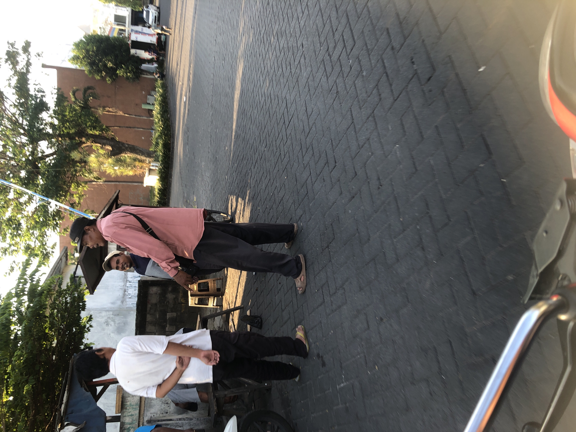 In Boro, Indonesia, under a clear sky with sunlight casting long shadows, three men are engaged in conversation on a paved street. The man on the right, wearing a pink long-sleeved shirt and a black cap, gestures while speaking. To his left, a man in a gray and blue shirt looks towards the speaker. The third man, wearing a white polo shirt and black pants, stands with his back to the camera, his hands clasped behind him. A motorcycle, partially visible in the foreground, suggests a street-level perspective. In the background, trees, buildings, and parked vehicles contribute to the urban setting.