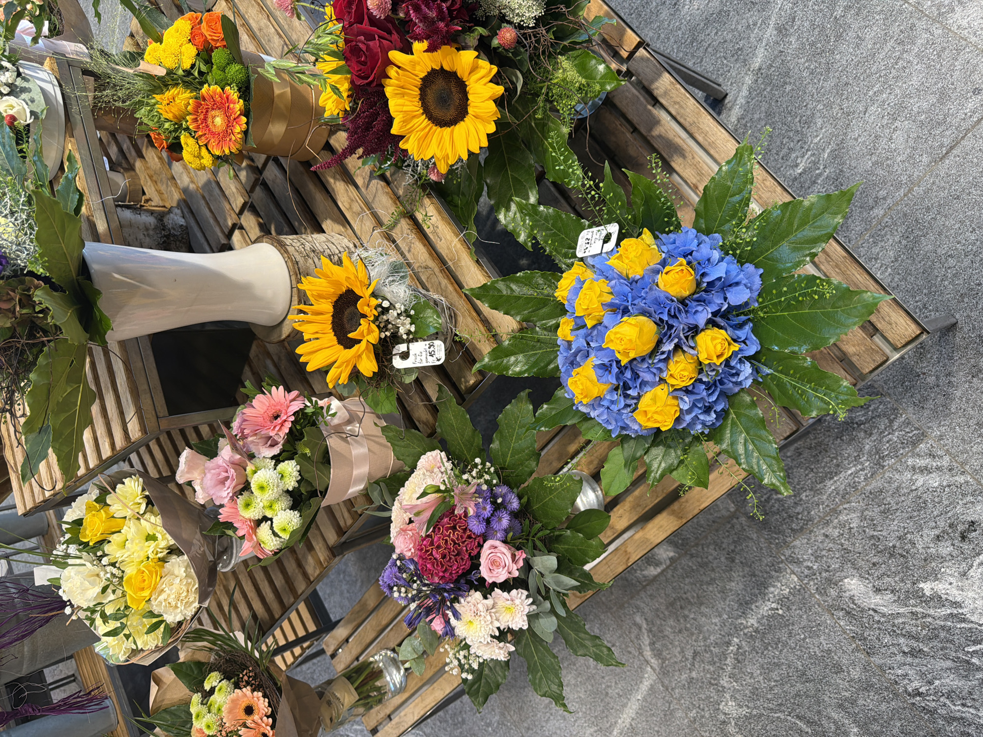 The image displays a collection of flower arrangements on wooden display shelves. The flowers vary in color and type, including sunflowers, roses, gerbera daisies, hydrangeas, and chrysanthemums. Some arrangements are in vases, while others are presented in baskets or wrapped bouquets. The scene appears to be an outdoor setting, possibly a market or flower shop, with grey tiled flooring visible in the foreground. There are no people or animals in the image. The lighting suggests it is daytime.