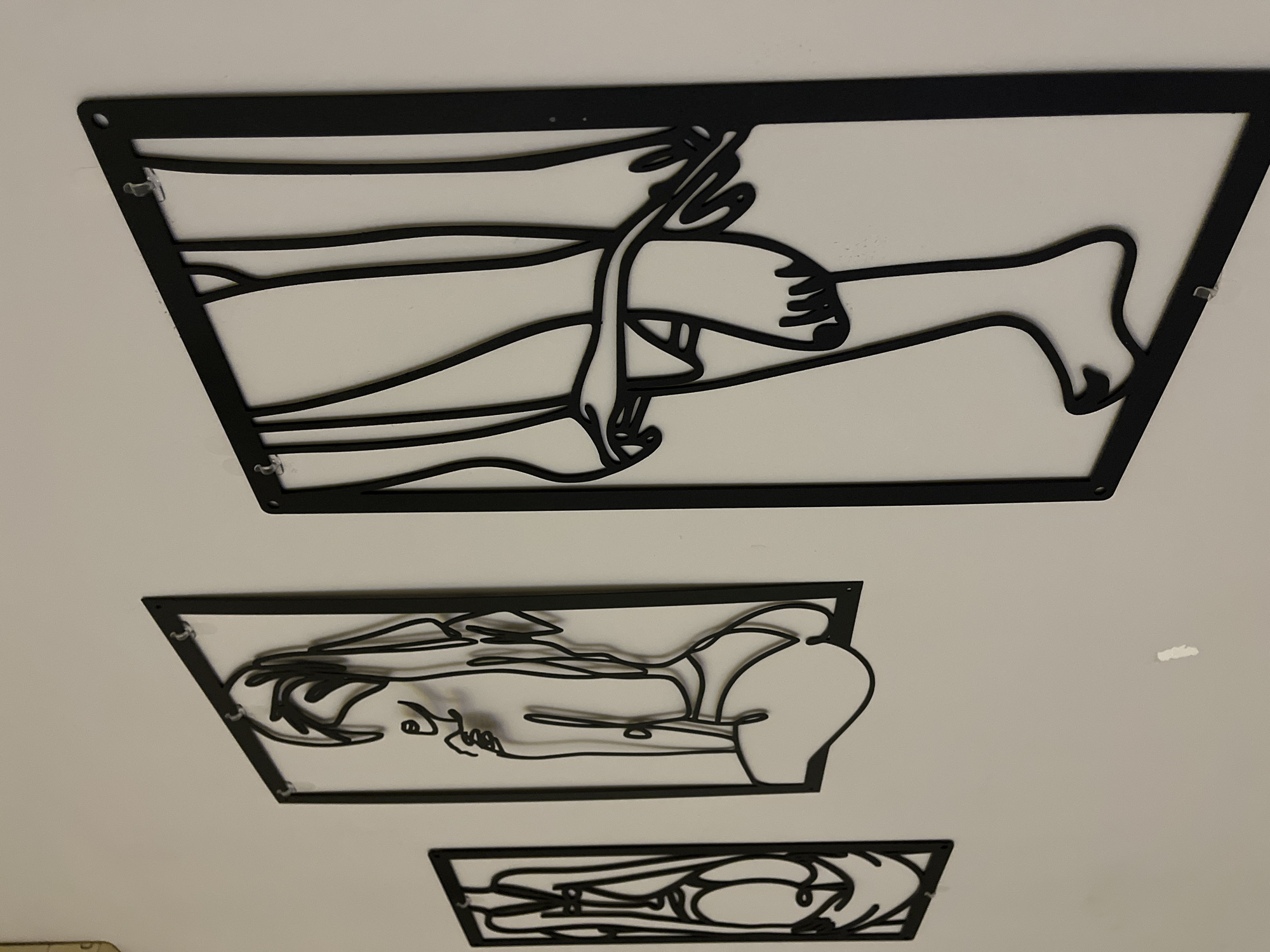 Three black metal cutout artworks hang on a white wall. The top artwork depicts intertwined arms, legs, and a foot. The middle artwork shows a reclining person with a head, chest, and buttocks. The bottom artwork features an ambiguous abstract form. Small clear plastic fasteners attach the artworks to the wall. The scene could be located in Veliko Tarnovo, Bulgaria.