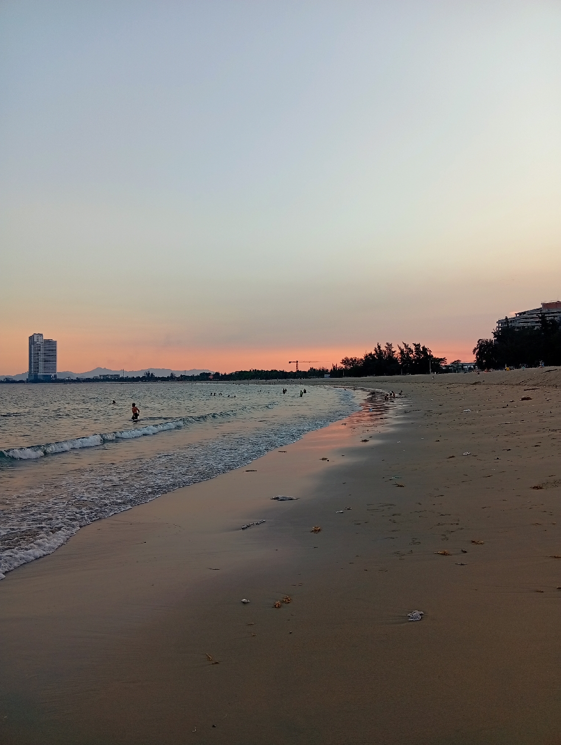 A wide-angle photograph captures a tranquil beach scene in Khánh Hải, Vietnam, during twilight, most likely sunset. The sky displays a beautiful gradient, transitioning from a soft light blue-grey at the top to warm orange and pink hues along the horizon.
In the far distance on the left, a tall, modern high-rise building with a light facade stands prominently against the colorful sky. A faint outline of distant mountains or hills is visible along the horizon line. Towards the mid-right, a line of dense trees separates the beach from an area with more buildings, possibly resort structures, and a construction crane is visible above the treetops.
The left side of the image features the ocean, with gentle waves breaking onto the sandy shore. Several people are visible in the water, enjoying a swim or wading, with one individual clearly standing in the shallow surf in the mid-left. More figures are scattered further along the beach and in the water, suggesting a popular spot.
The foreground is dominated by a wide stretch of light-colored sand, showing the imprint of receding waves and scattered with what appears to be natural debris like shells or small pieces of wood. The overall lighting is soft and warm, creating a peaceful and serene atmosphere. No visible text is present in the image.