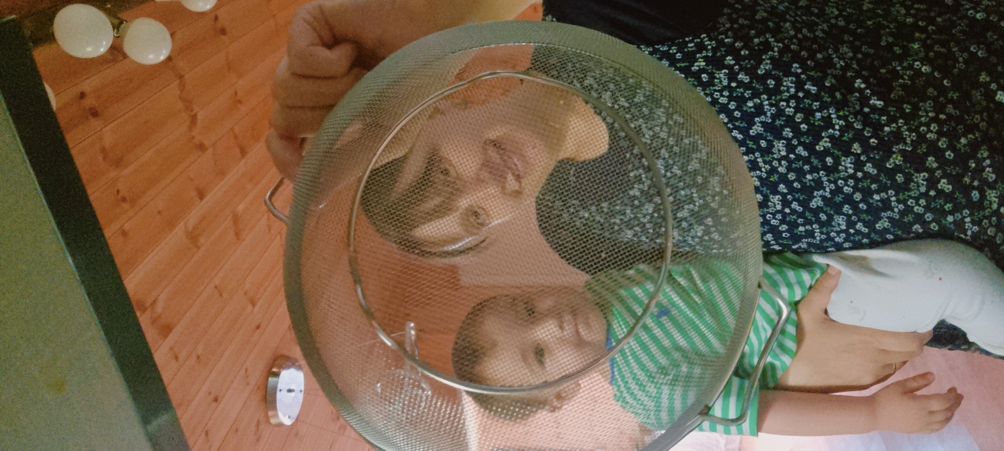 A colander is held over two upside-down infants and one adult, likely indoors in Dunnae-myeon, South Korea. The infants wear a green and white striped shirt and an orange shirt. The adult is holding a pillow. The environment shows a wooden floor and a countertop. The colander is made of metal.